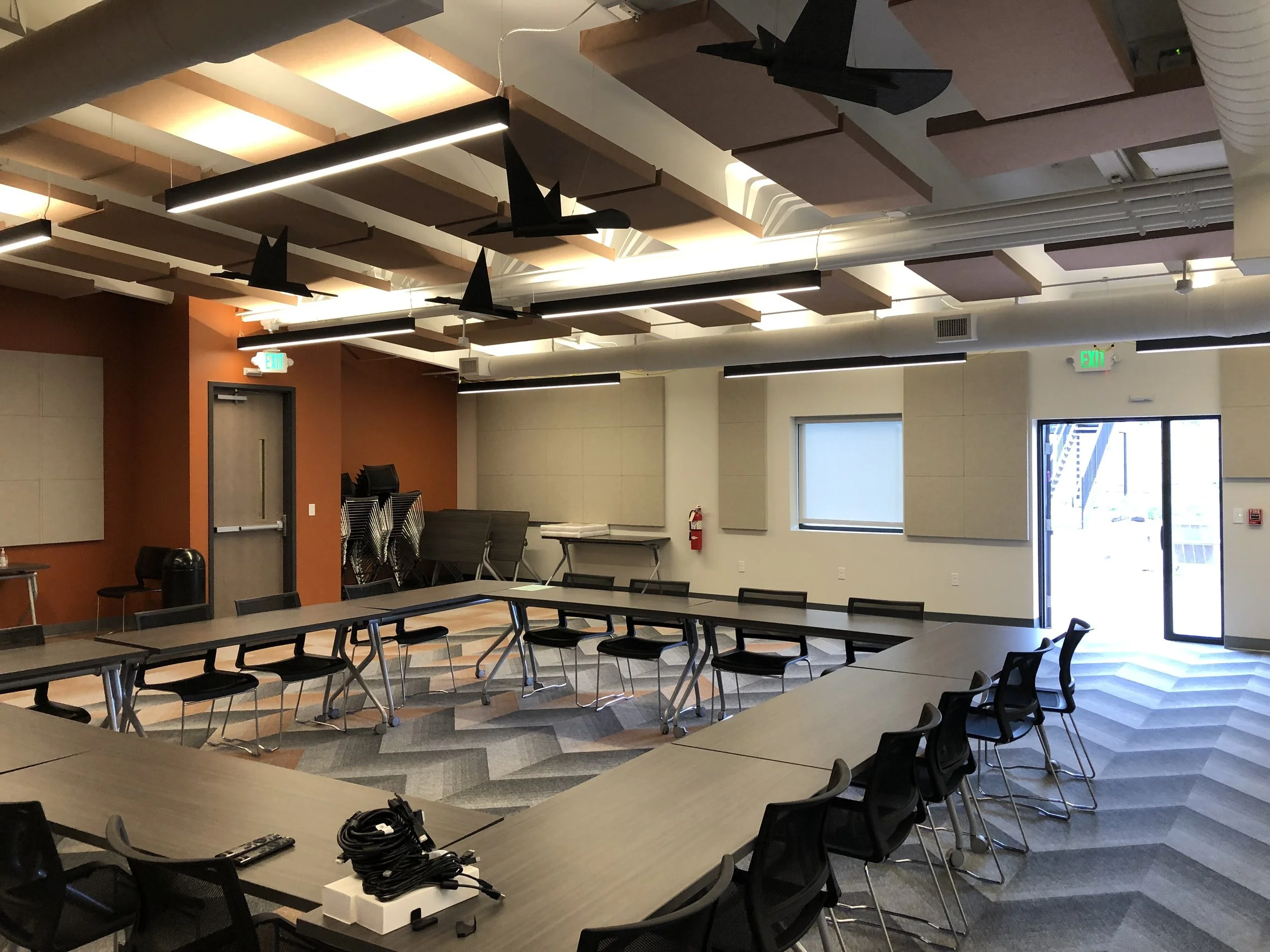 A modern conference room with U-shaped tables and black chairs, featuring geometric carpet patterns. The room has acoustic panels on the ceiling and walls, and a stack of chairs and tables in the corner. There's a door with exit signs and a visible fire extinguisher.