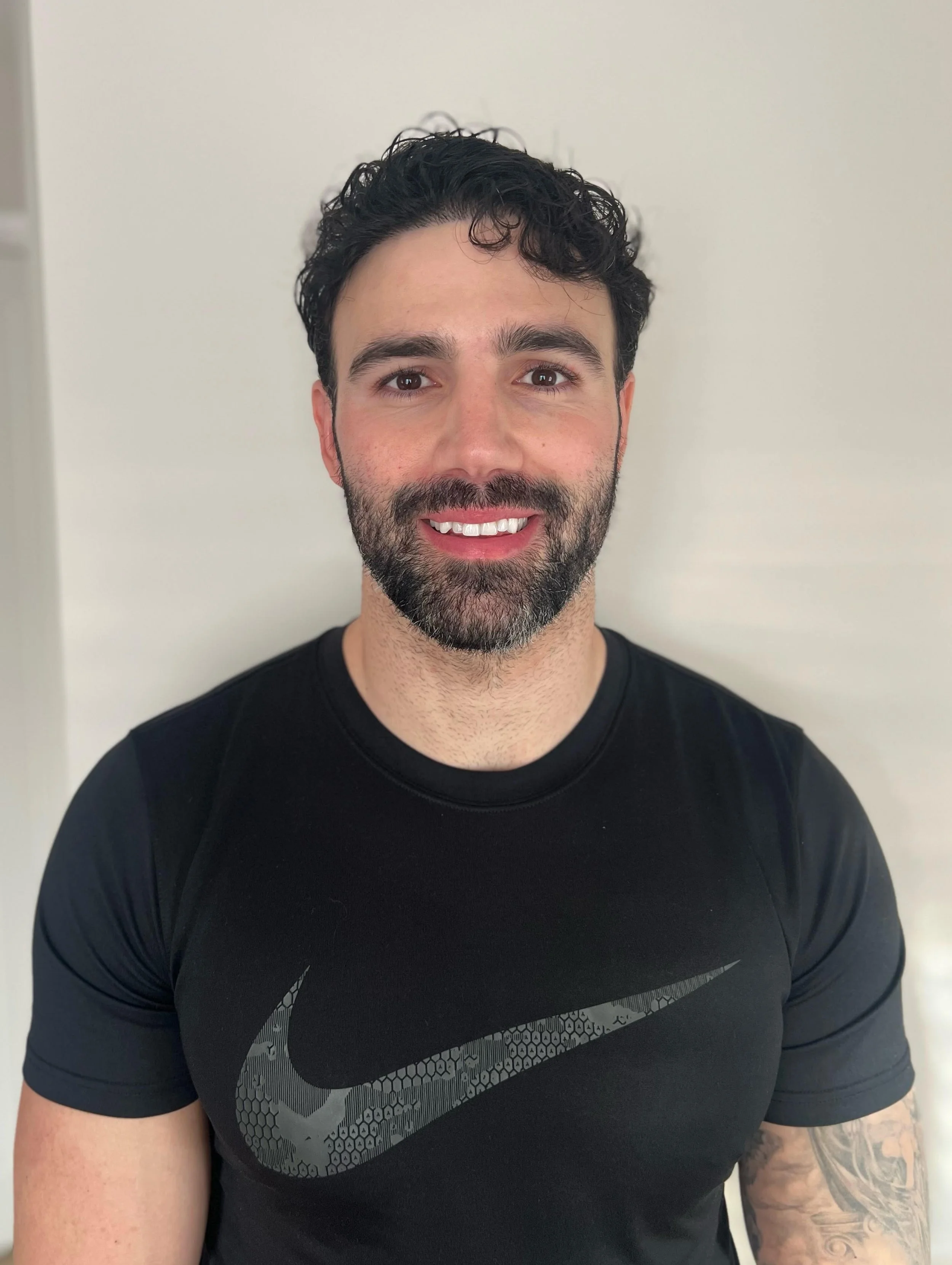 A smiling man with dark curly hair and a beard, wearing a black Nike t-shirt, standing against a plain light-colored background.