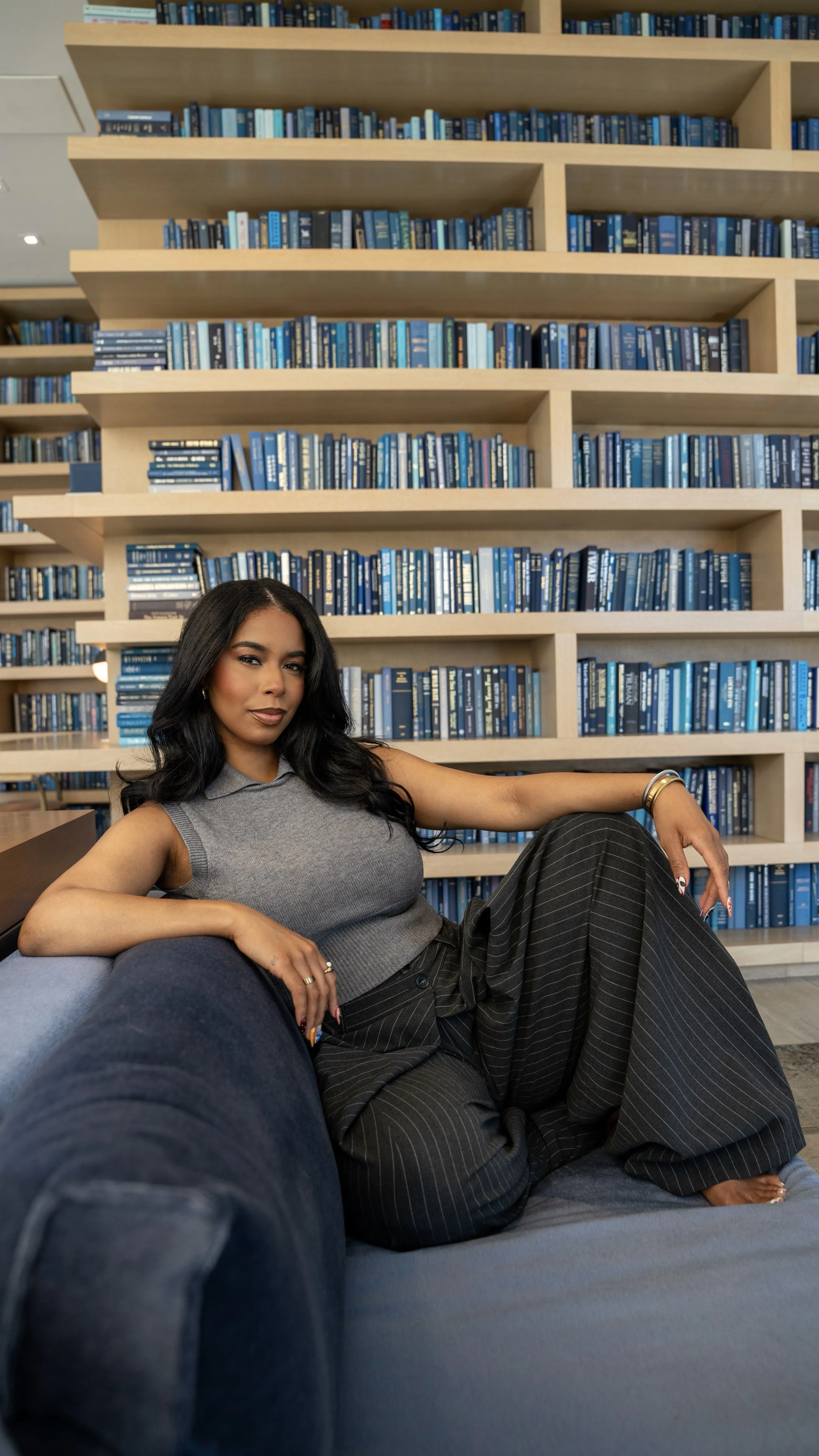 A woman lounging on a gray sofa in a library or bookstore with wooden shelves filled with blue books in the background.