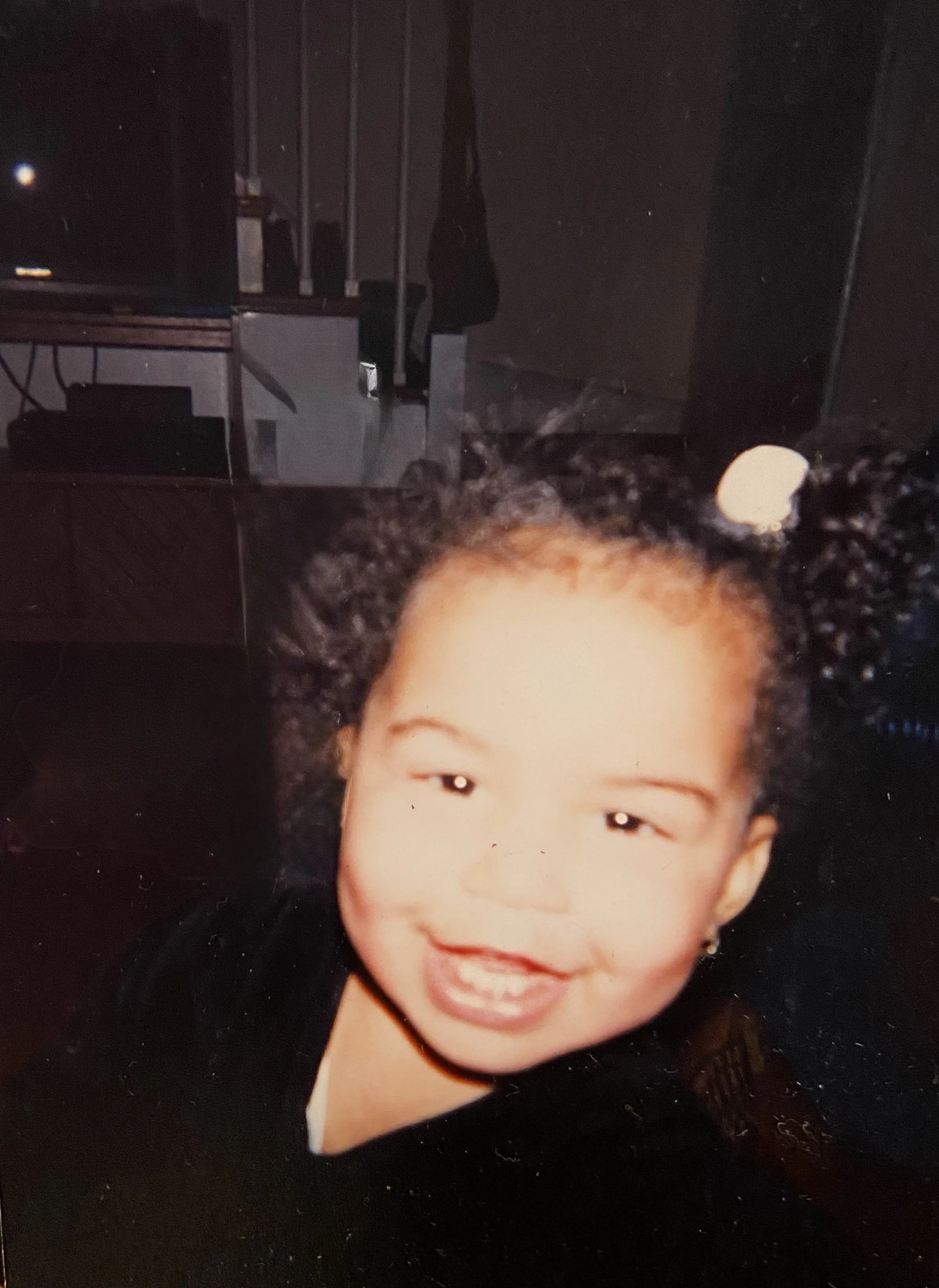 A young girl with curly black hair smiling in a dimly lit room.