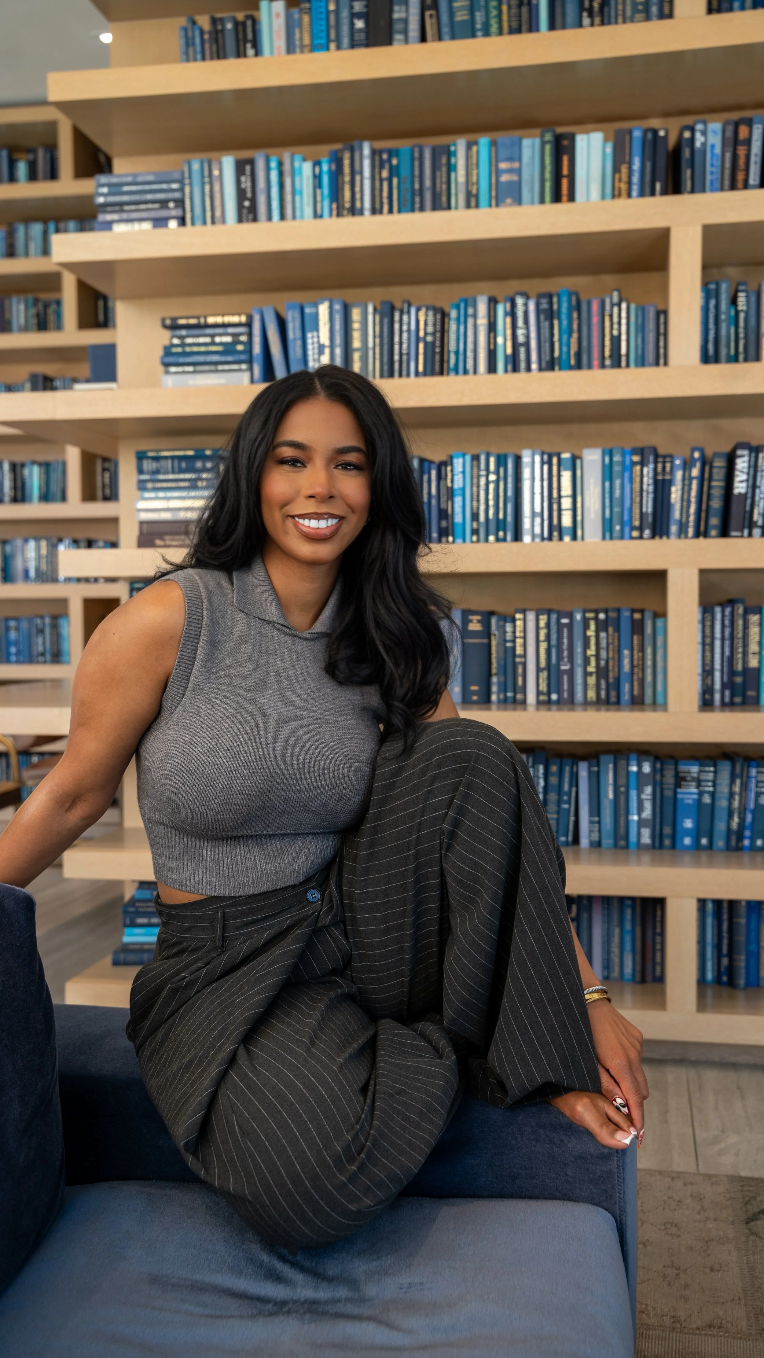 A smiling woman with long black hair, wearing a gray sleeveless top and black pinstripe pants, sitting on a blue sofa in front of a bookshelf filled with blue books.