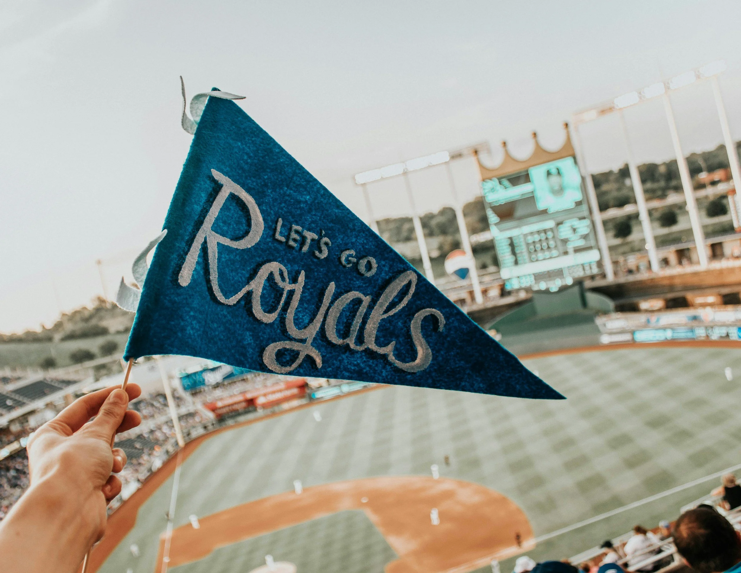 Royal blue pennant flag that reads 'Let's Go Royals' being held on a stick in front of the Crown shaped scoreboard featured in a blog called Birthday Gift For Husband in Kansas City.