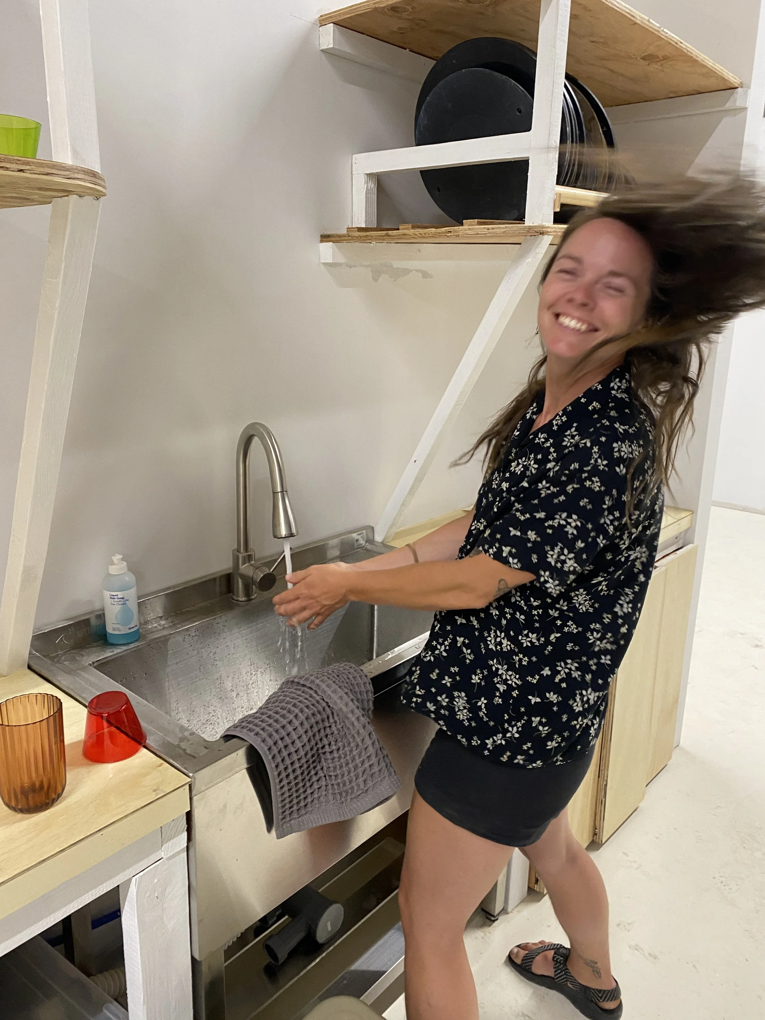 A woman with long brown hair, smiling, washing her hands at a kitchen sink.