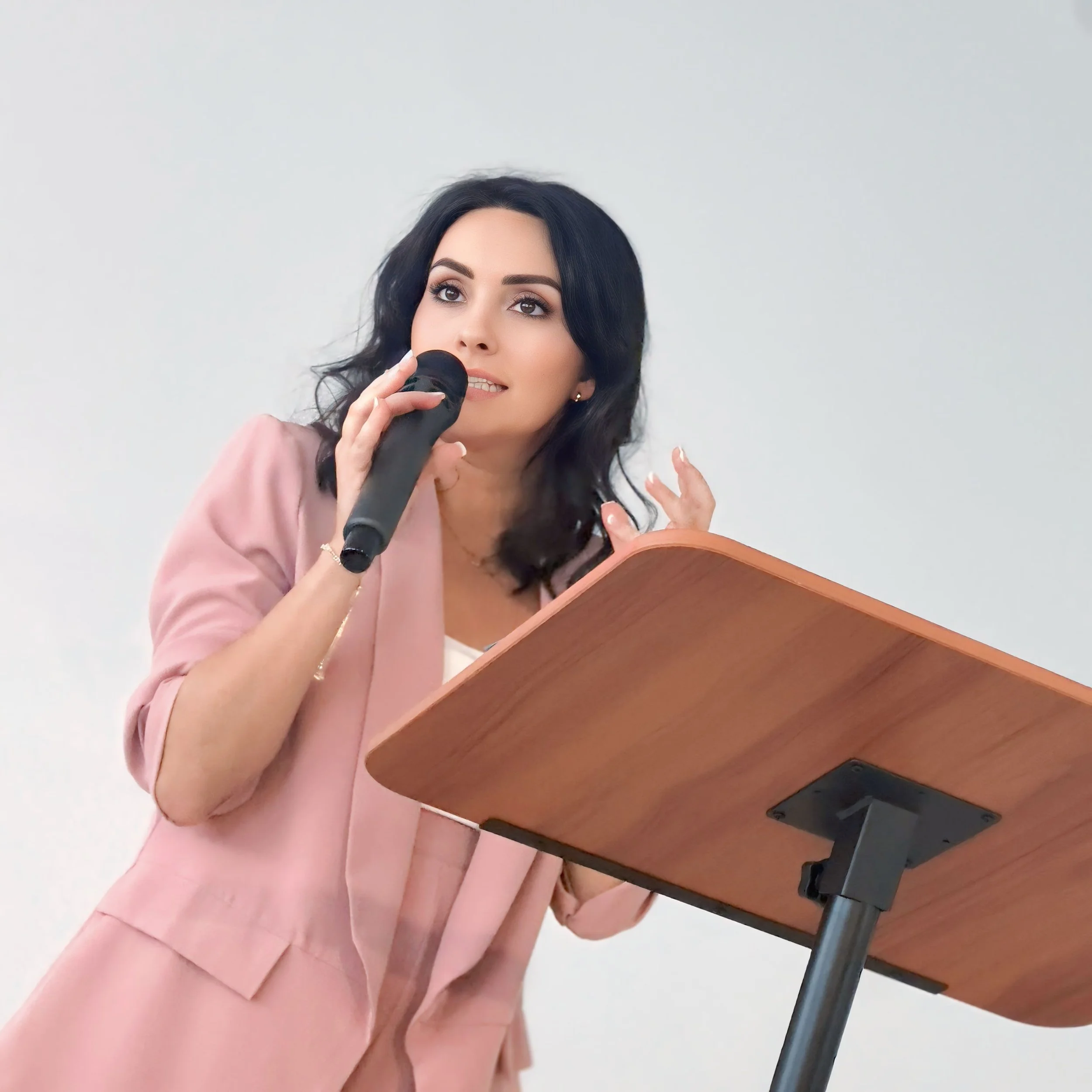 Woman speaking at a podium, holding a microphone with a plain white wall in the background.