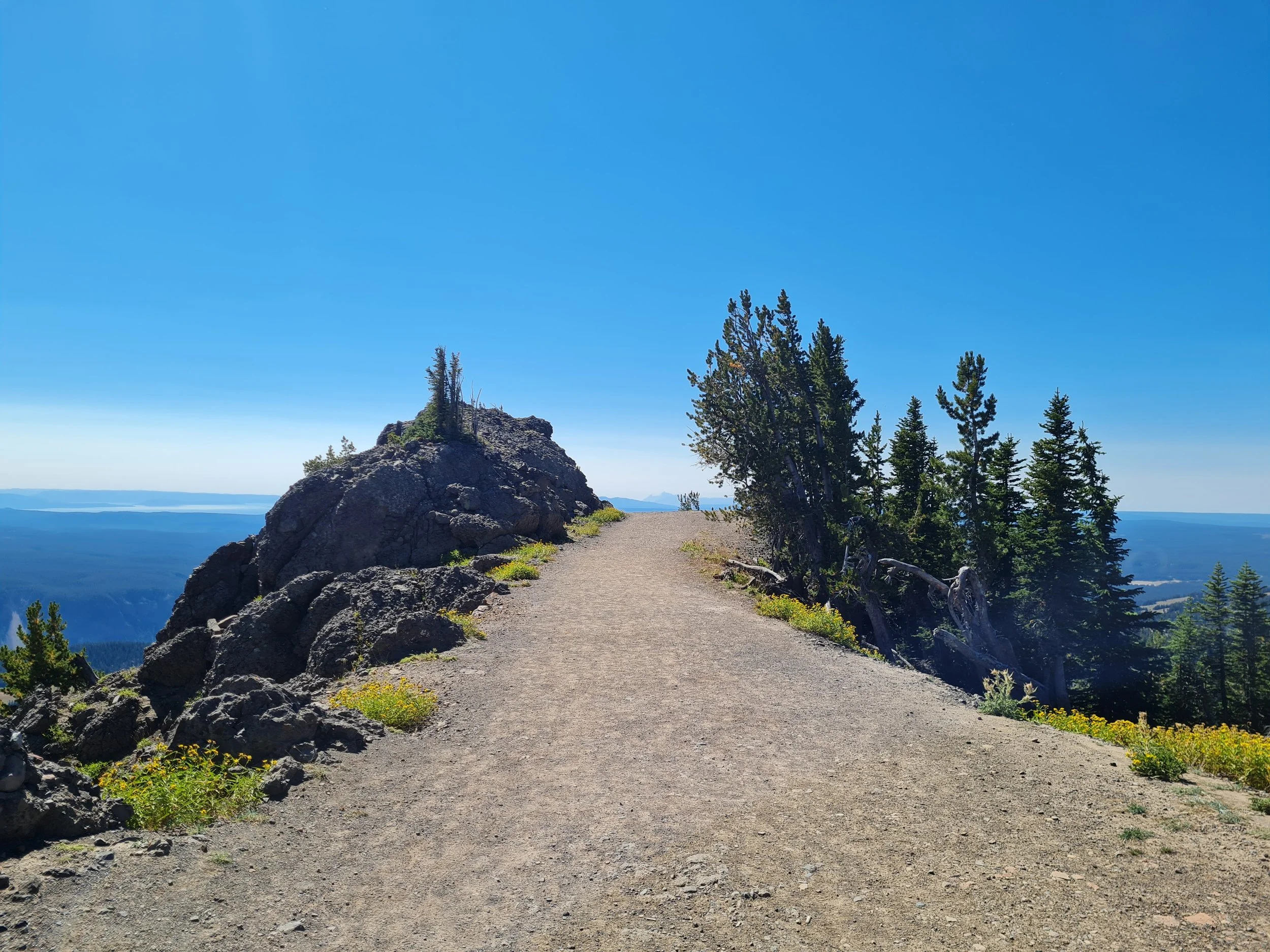 A dirt trail on a mountain ridge with large rocks and pine trees, overlooking a vast landscape of mountains and valleys under a clear blue sky.