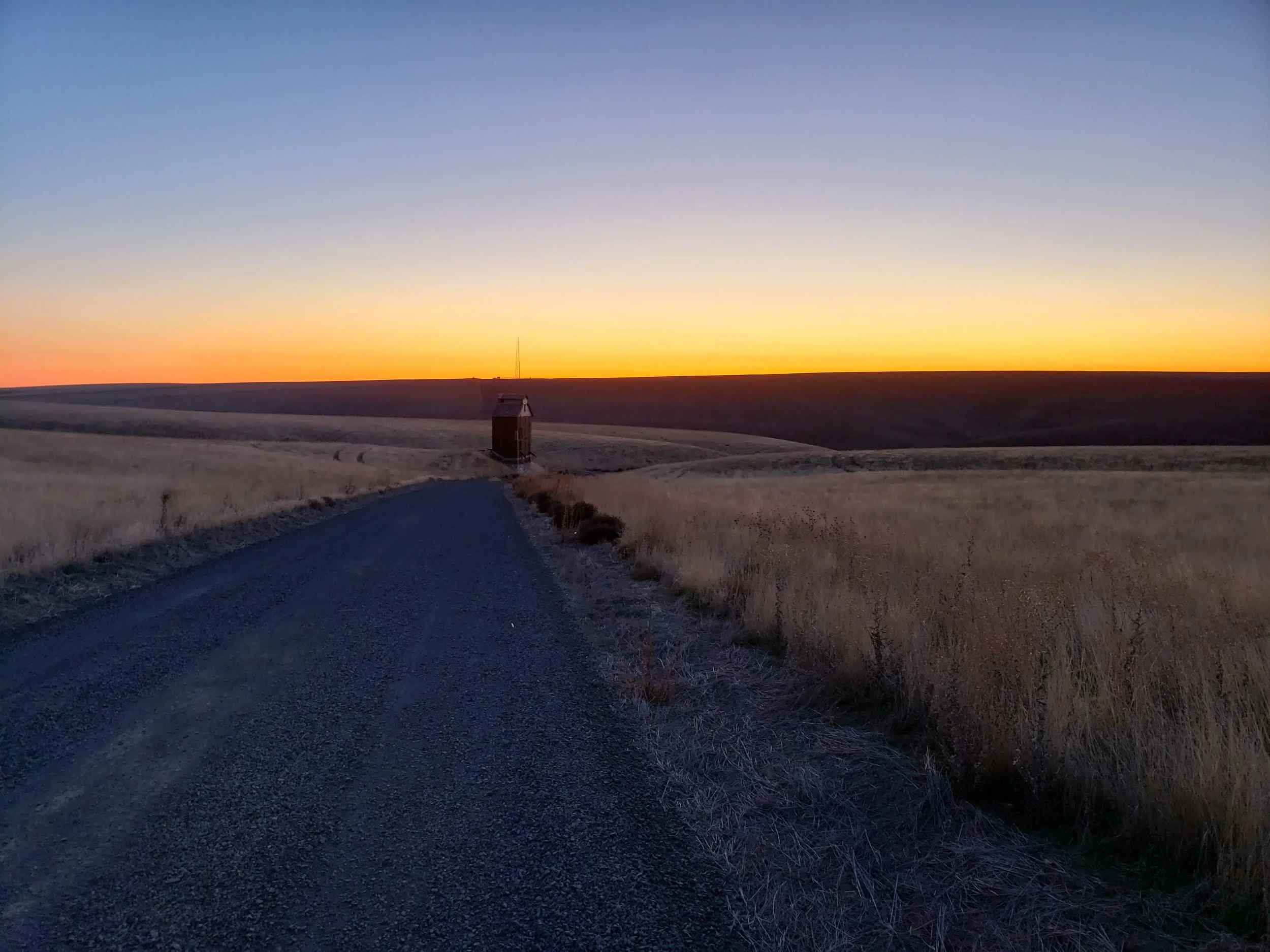 A gravel road leading towards a small wooden house with a tall antenna in a wide open field during sunset.