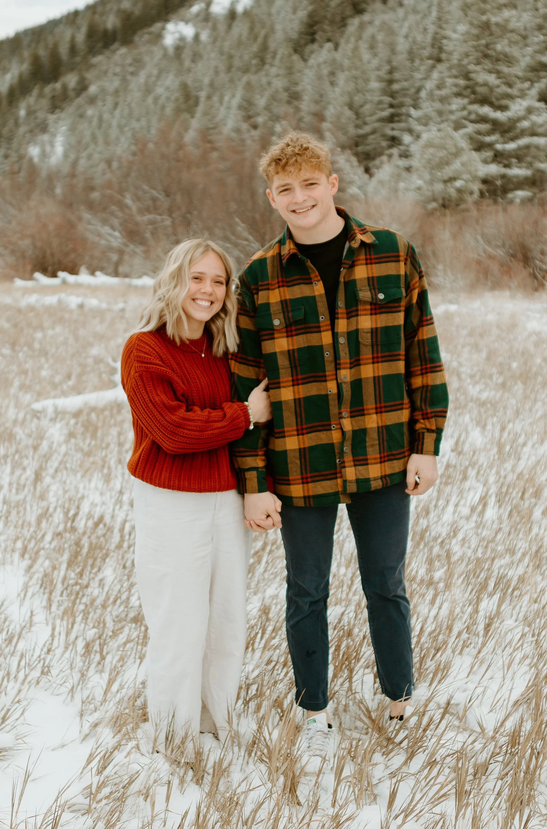 A smiling young woman and a young man standing in a snowy field, holding hands, with a forested hillside in the background.