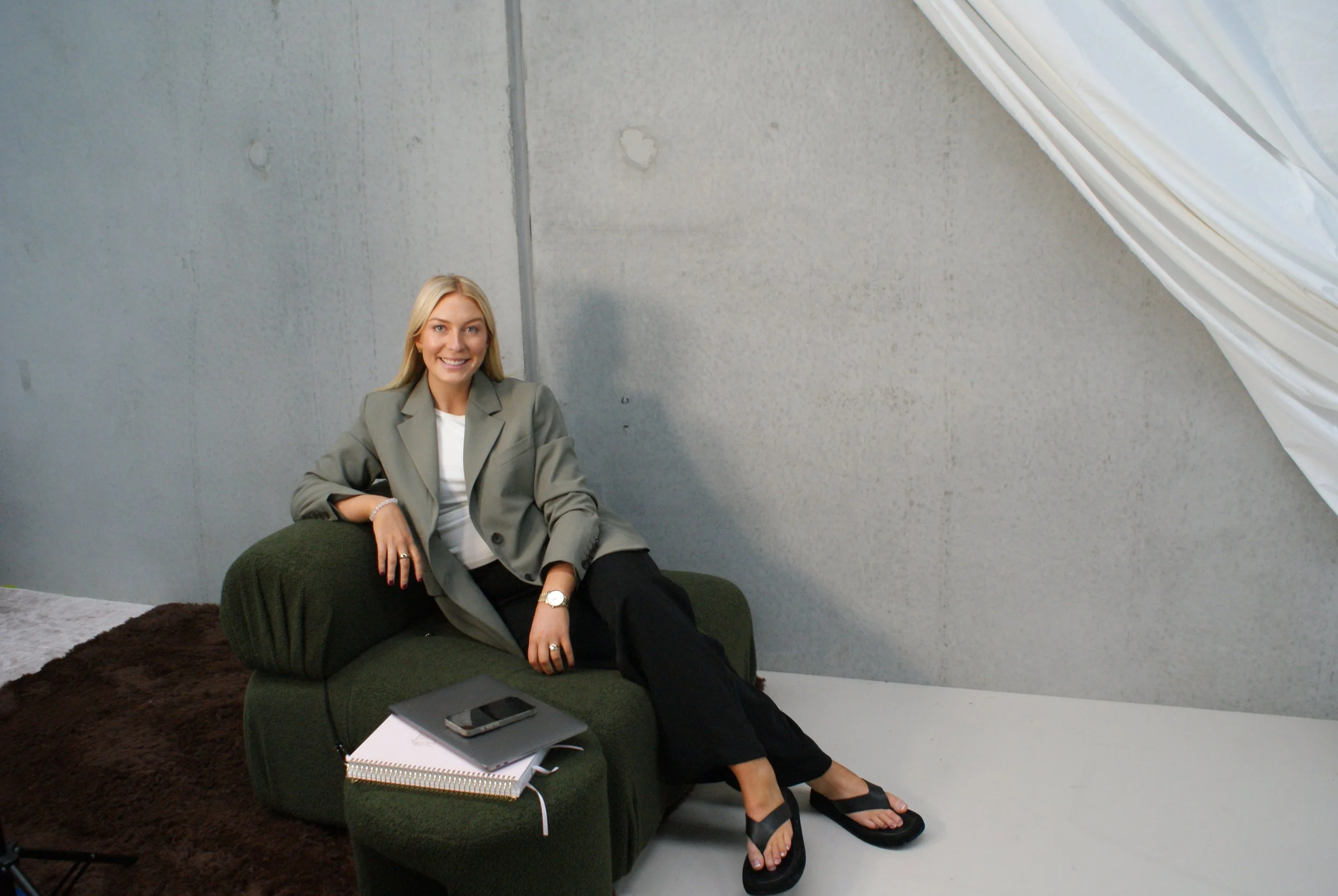 A young woman with blonde hair sitting on a green armchair, smiling, wearing a light gray blazer, black pants, and black flip-flops. There are notebooks, a phone, and a pen on the armchair.