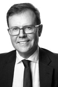 Black and white portrait of a man with glasses, short hair, wearing a suit and tie, smiling.