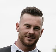 Portrait of a young man with short brown hair and a beard, wearing a tuxedo, smiling against a plain white background.