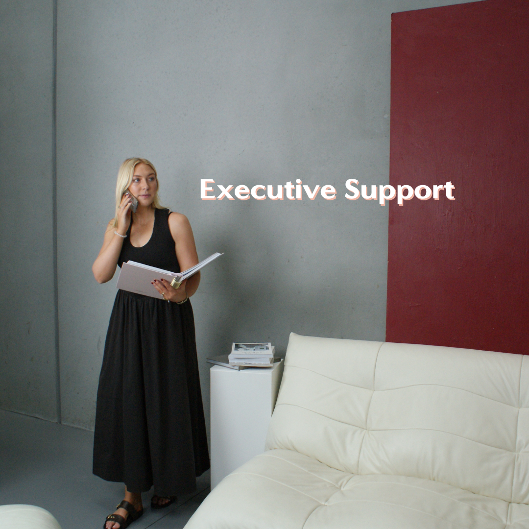 A woman with blonde hair in a black sleeveless dress holds a phone to her ear and a white folder, standing next to a white sofa in a modern office space. The wall behind her is gray with a red accent section, and there is a small white side table with stacks of papers or magazines. The text 'Executive Support' is overlaid on the image.
