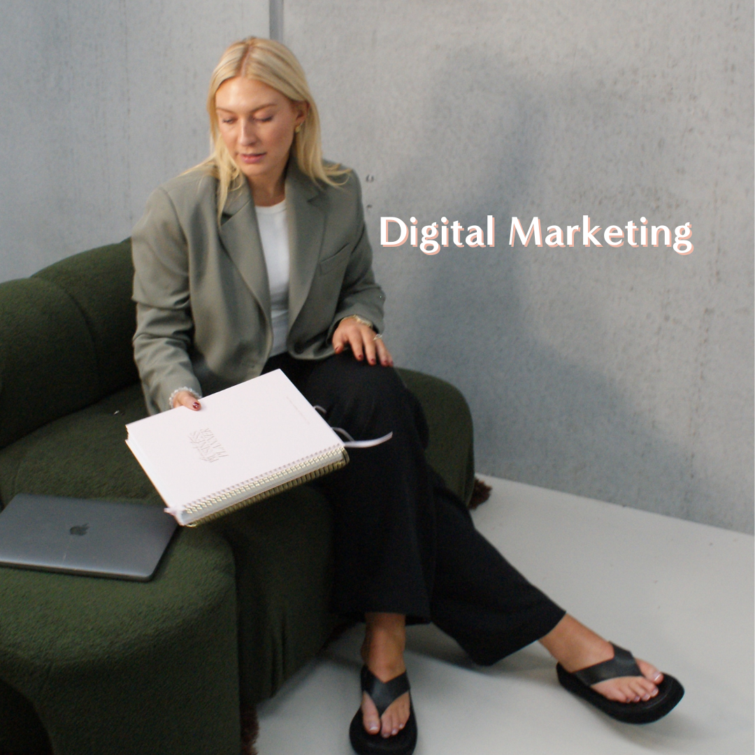 A woman sitting on a green couch with a laptop and notebook, looking at her notebook, with the text 'Digital Marketing' on the wall behind her.