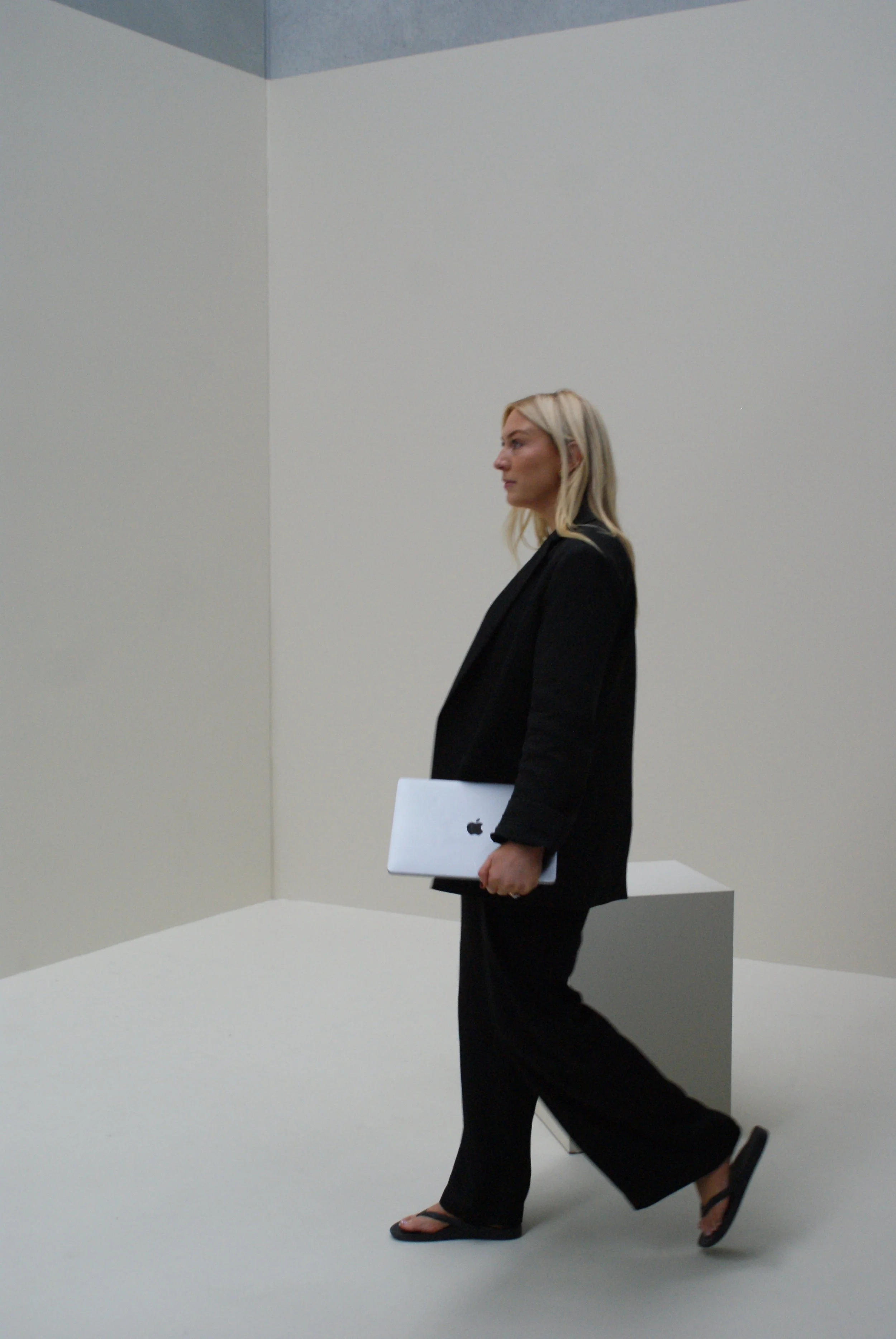 A woman with blonde hair wearing a black suit and sandals walking in a plain, minimalistic room and carrying a silver MacBook laptop.