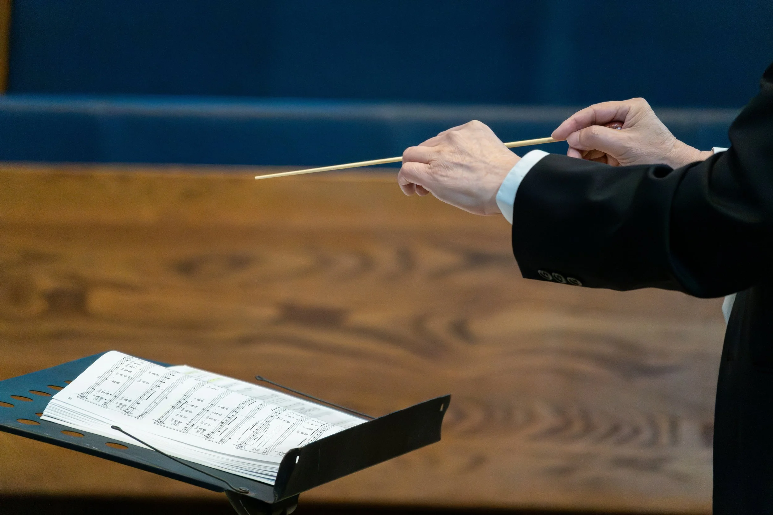The hands of a symphonic conductor in motion and holding a baton. A music stand with a musical score is on the left-hand side.