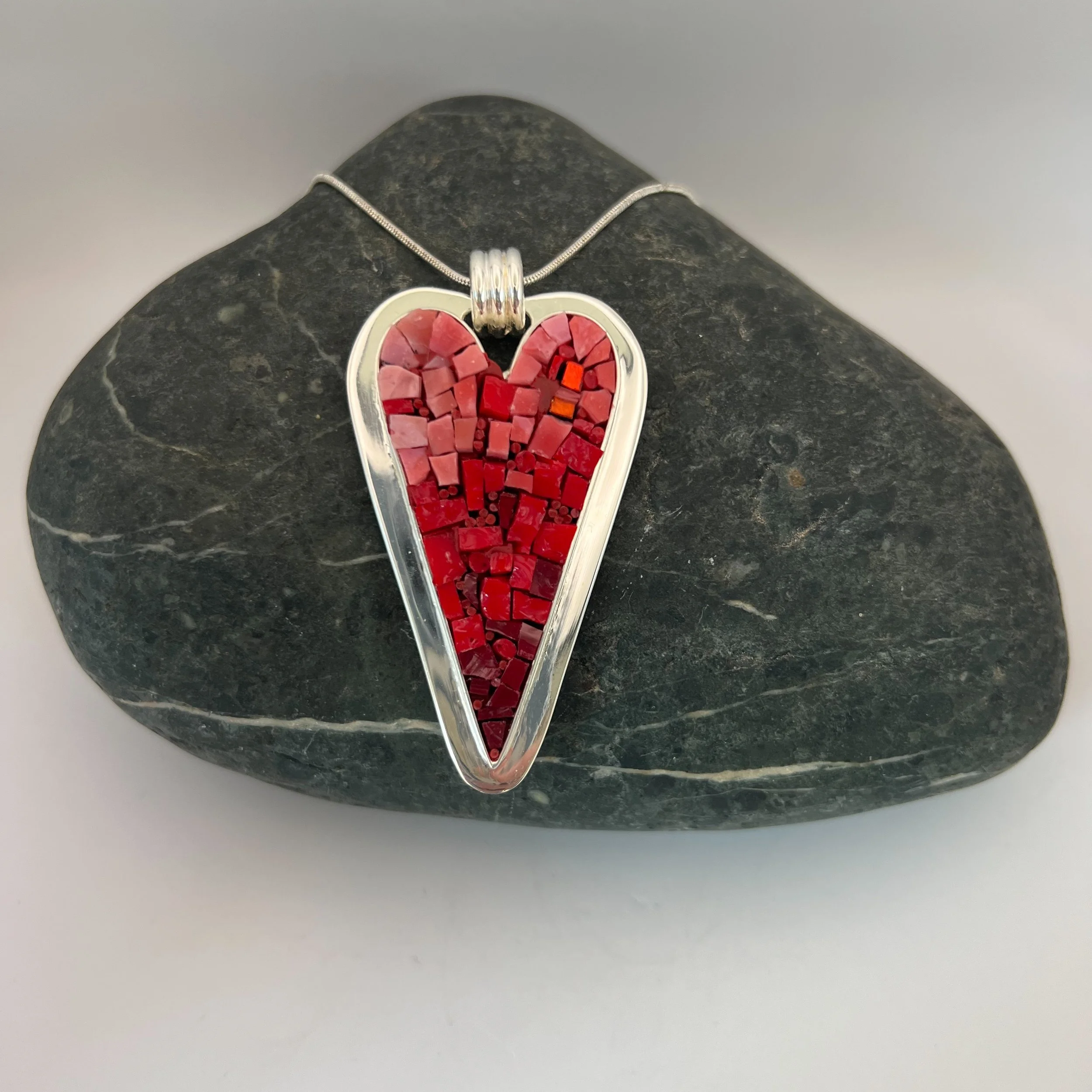 Heart shaped mosaic pendant with red glass drop in a silver frame placed on a gray stone heart-shaped surface.