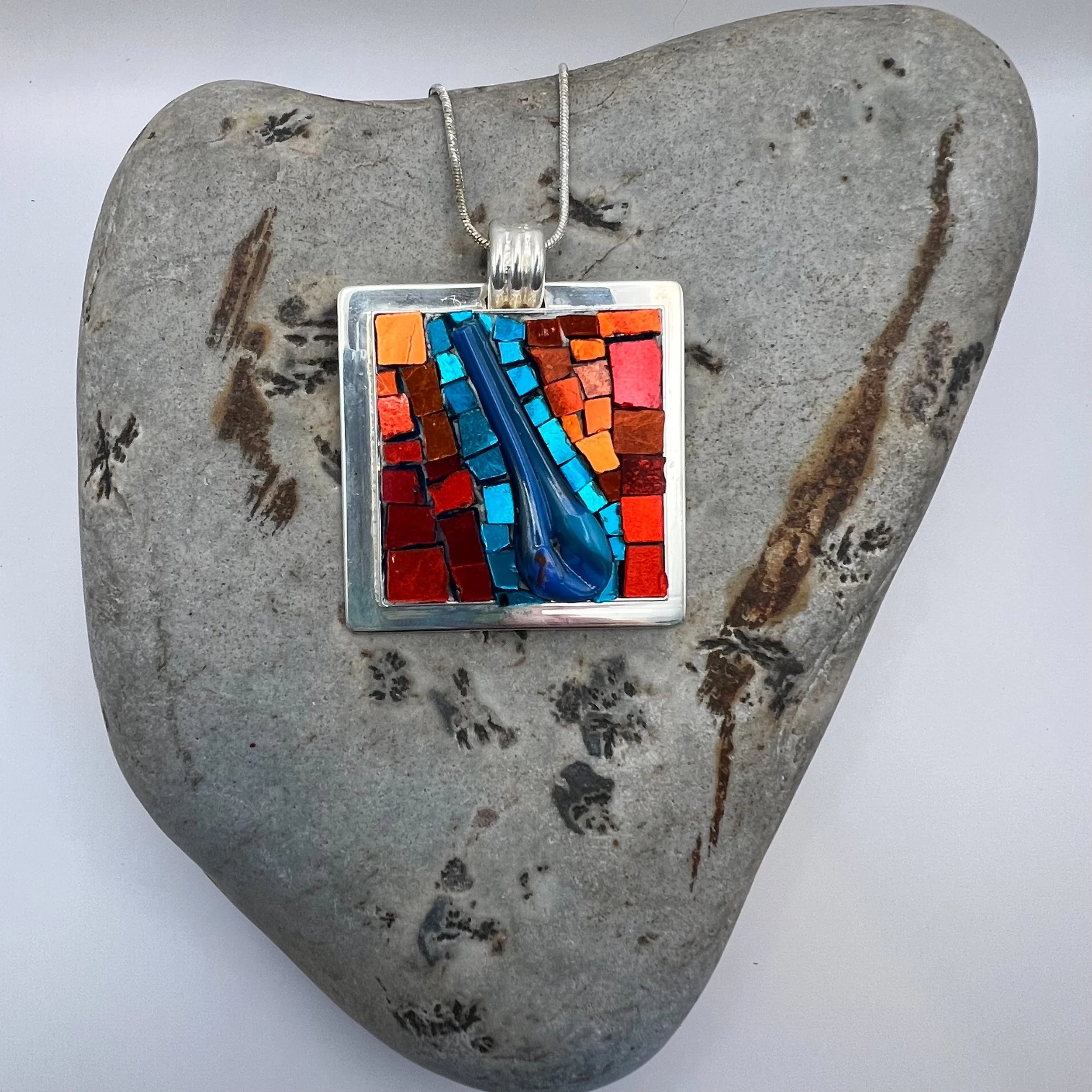 Colorful square mosaic pendant with blue glass drop in a silver frame placed on a gray stone heart-shaped surface.