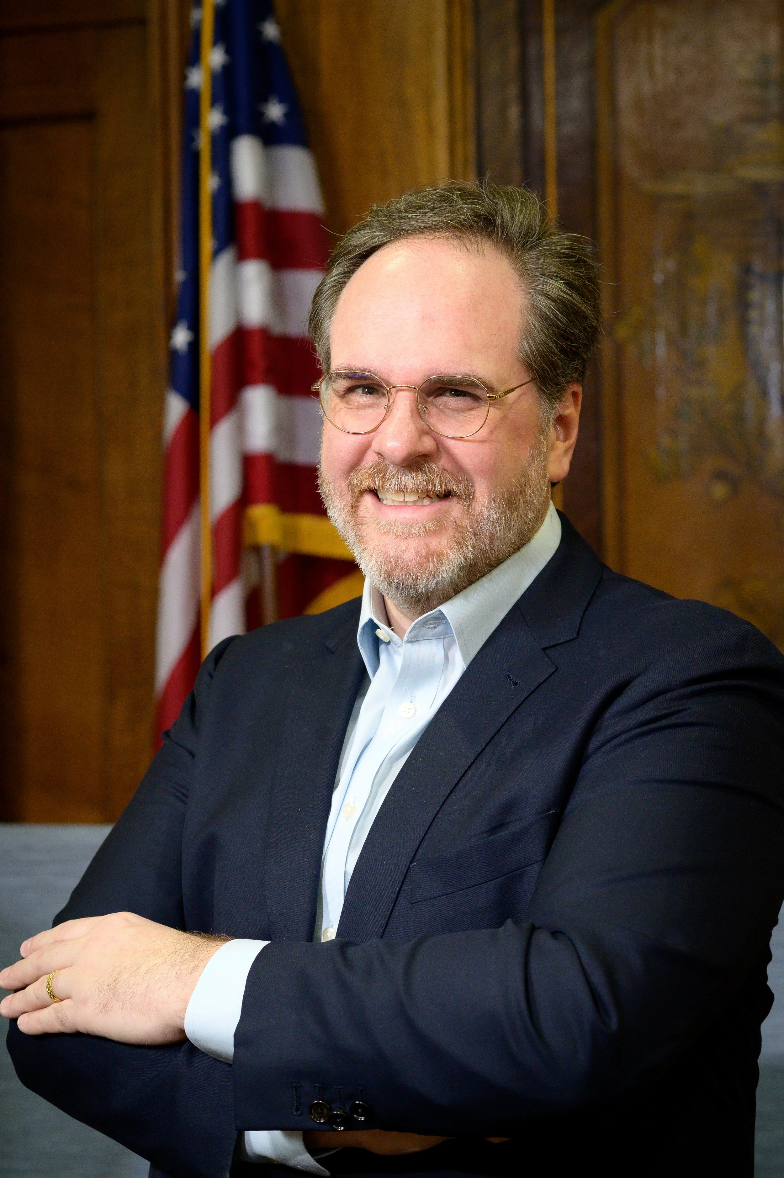 A man with glasses and a beard wearing a dark suit and light shirt, smiling, standing in front of a wood-paneled wall and a blurred American flag.