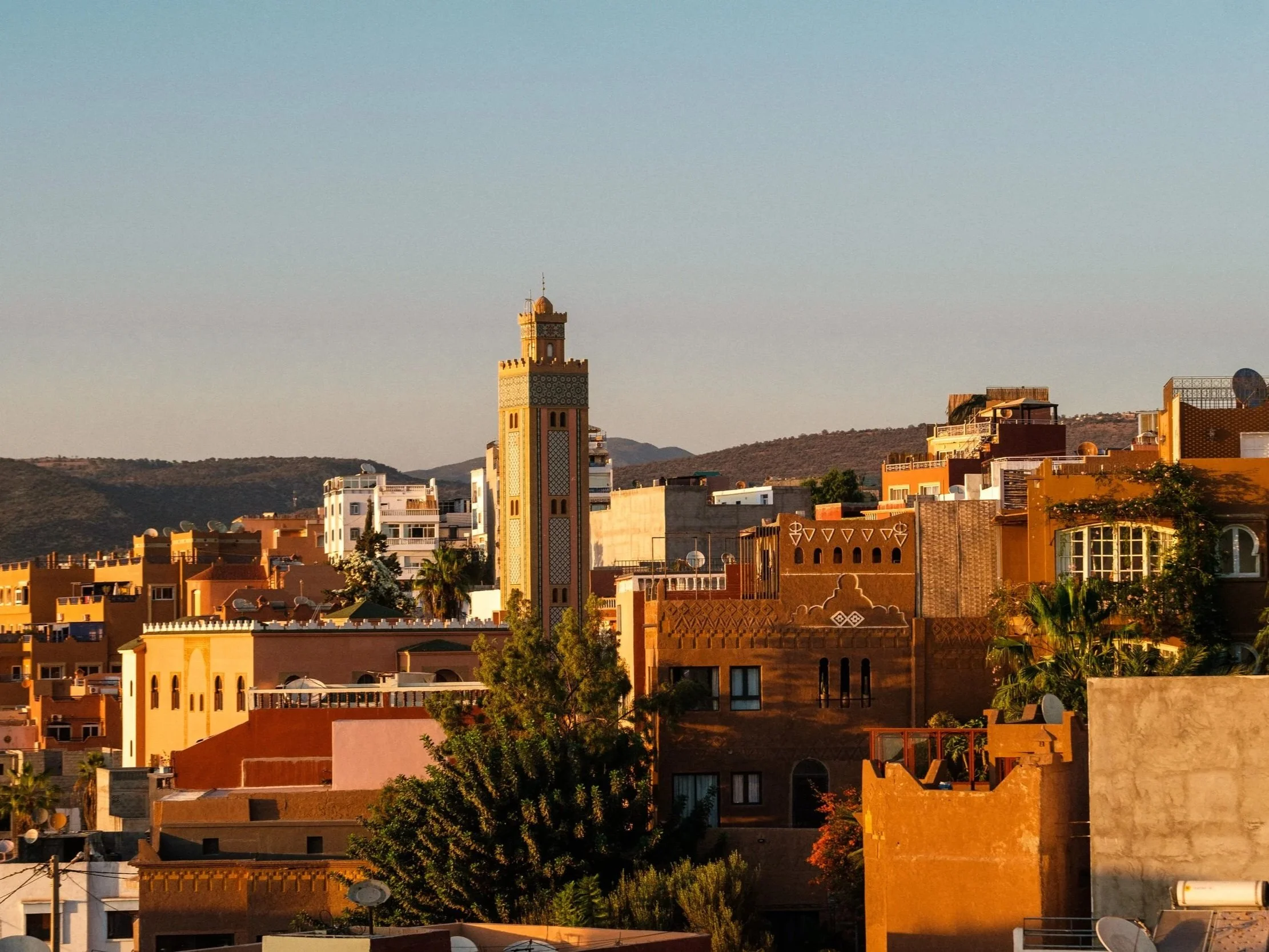 Sunset view of a cityscape with traditional and modern buildings, including a prominent beige minaret-style tower, with hills in the background.