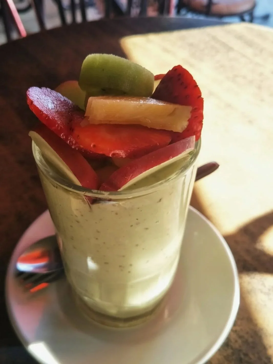 A glass of fruit smoothie topped with sliced strawberries, kiwi, pineapple, and apple, set on a white saucer with a spoon beside it on a wooden table.