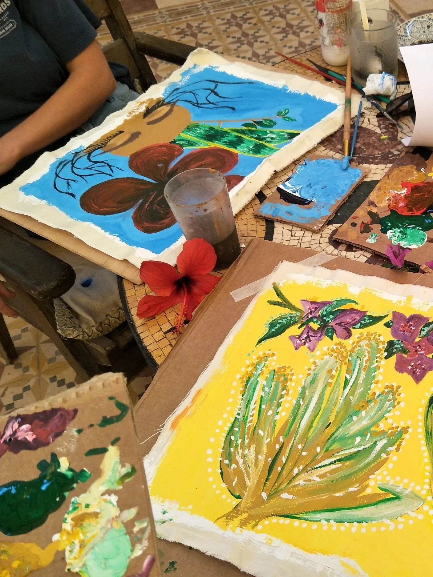 Paintings with floral and nature themes on paper, various brushes, paints, and a glass of water on a mosaic-tiled table.