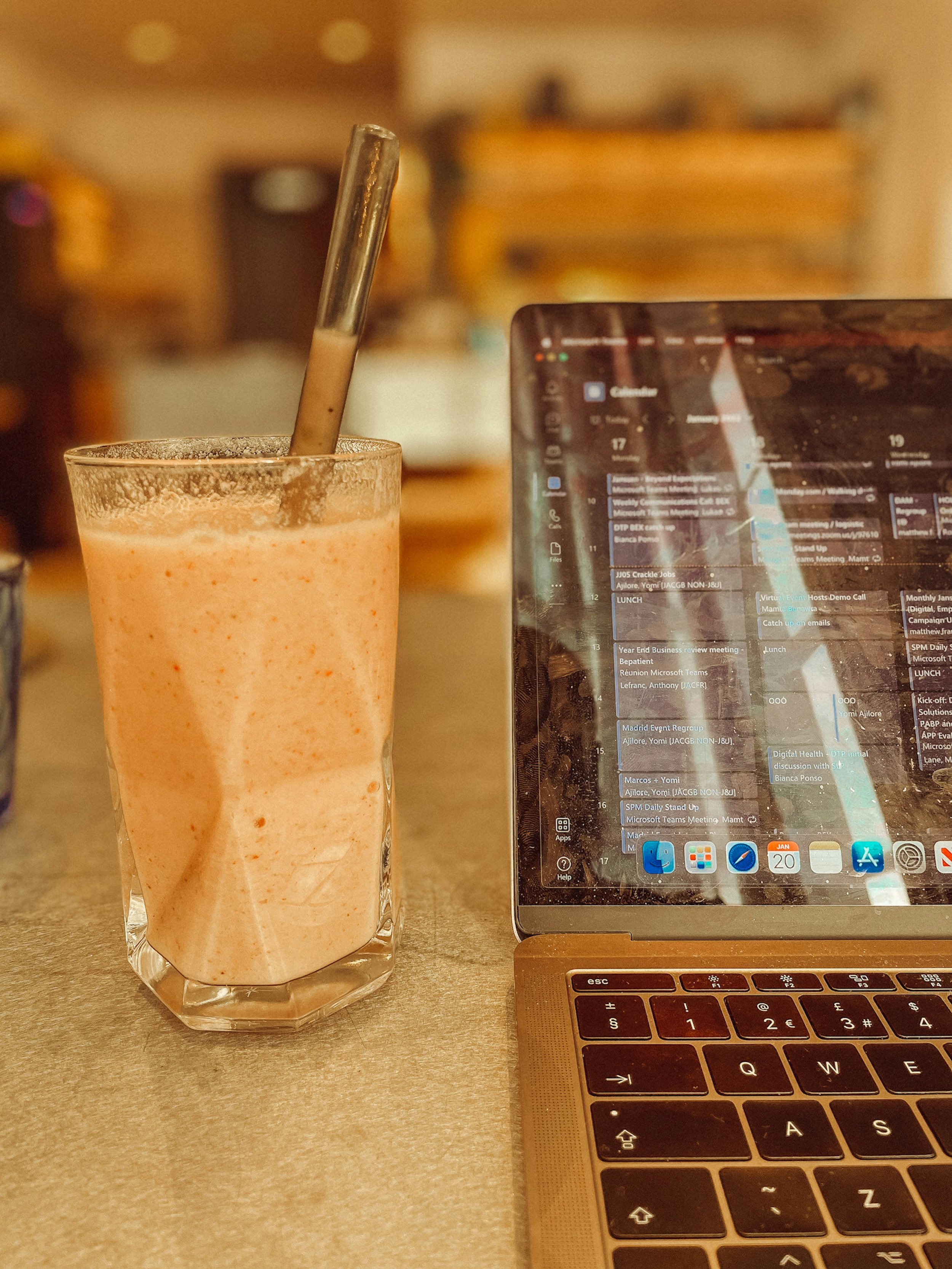 A glass of pink smoothie with a straw next to a laptop on a table.