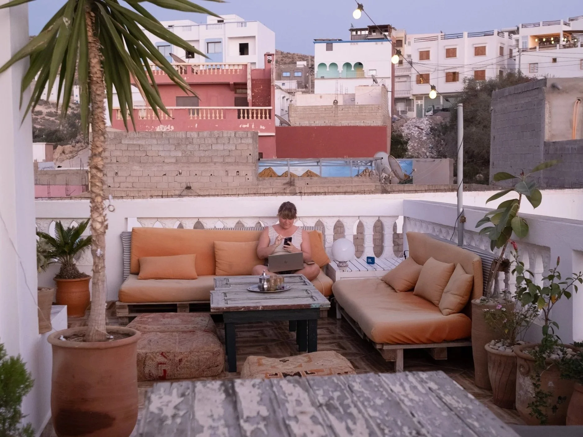 A woman sitting on an outdoor patio couch using her phone, surrounded by potted plants and outdoor furniture, with a cityscape of buildings in the background.