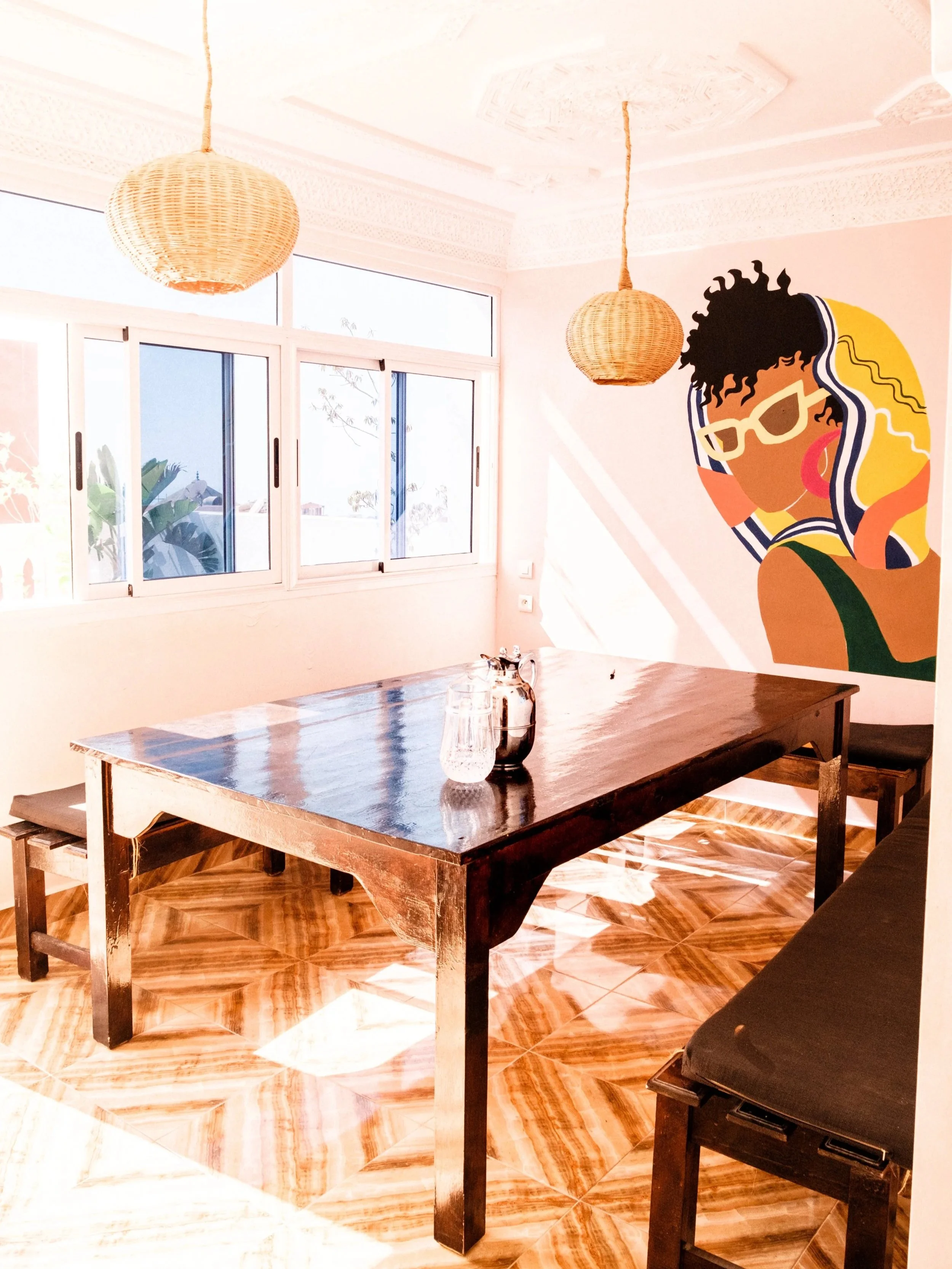 A dining room with a wooden table, black chairs, a pitcher, and a glass of water. There is a colorful mural of a woman with glasses on the wall, two woven pendant lights hanging from the ceiling, and a large window allowing natural light into the room.