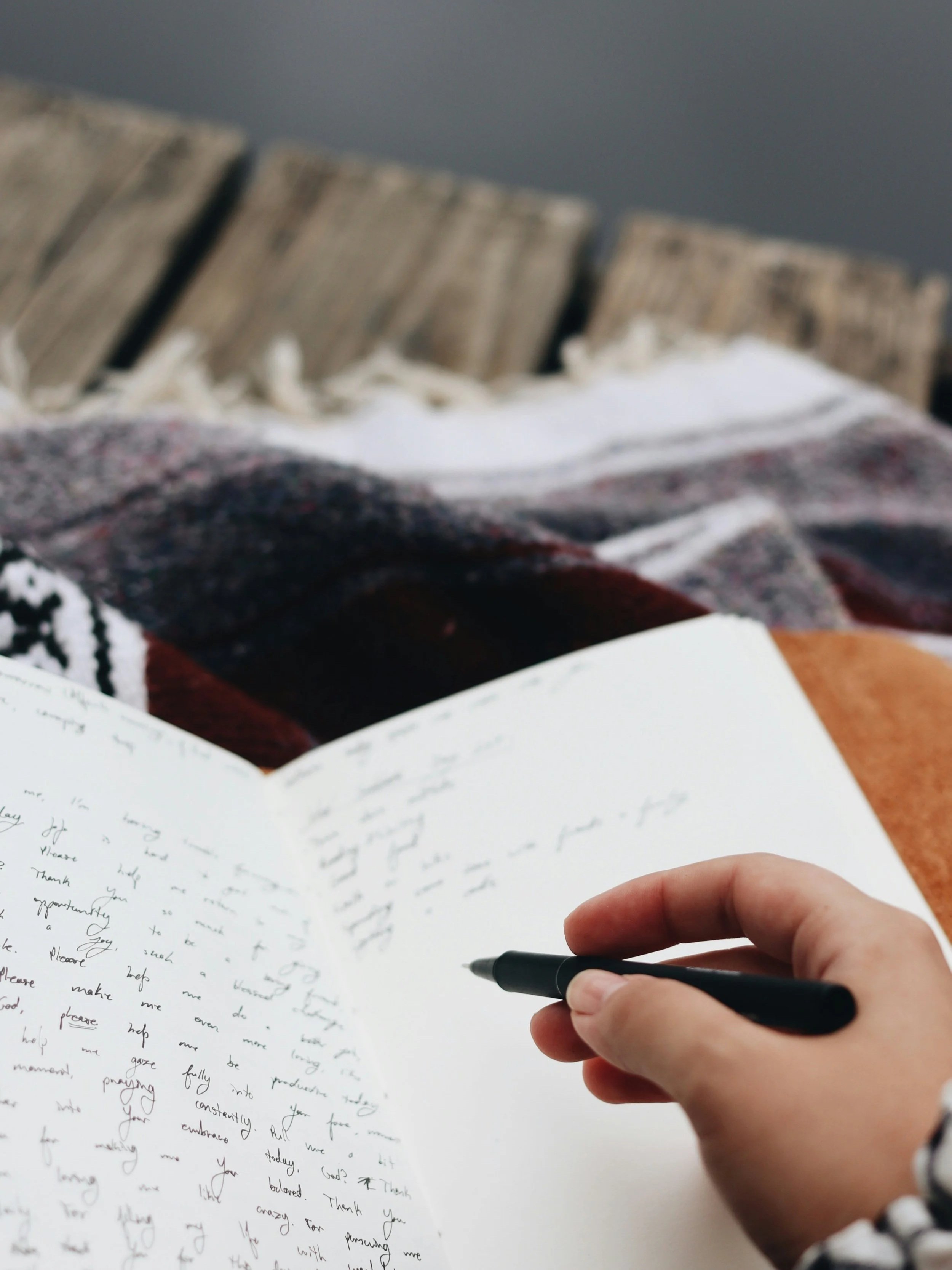 A person holding a pen and writing in a notebook, with a blanket and a wooden surface in the background.
