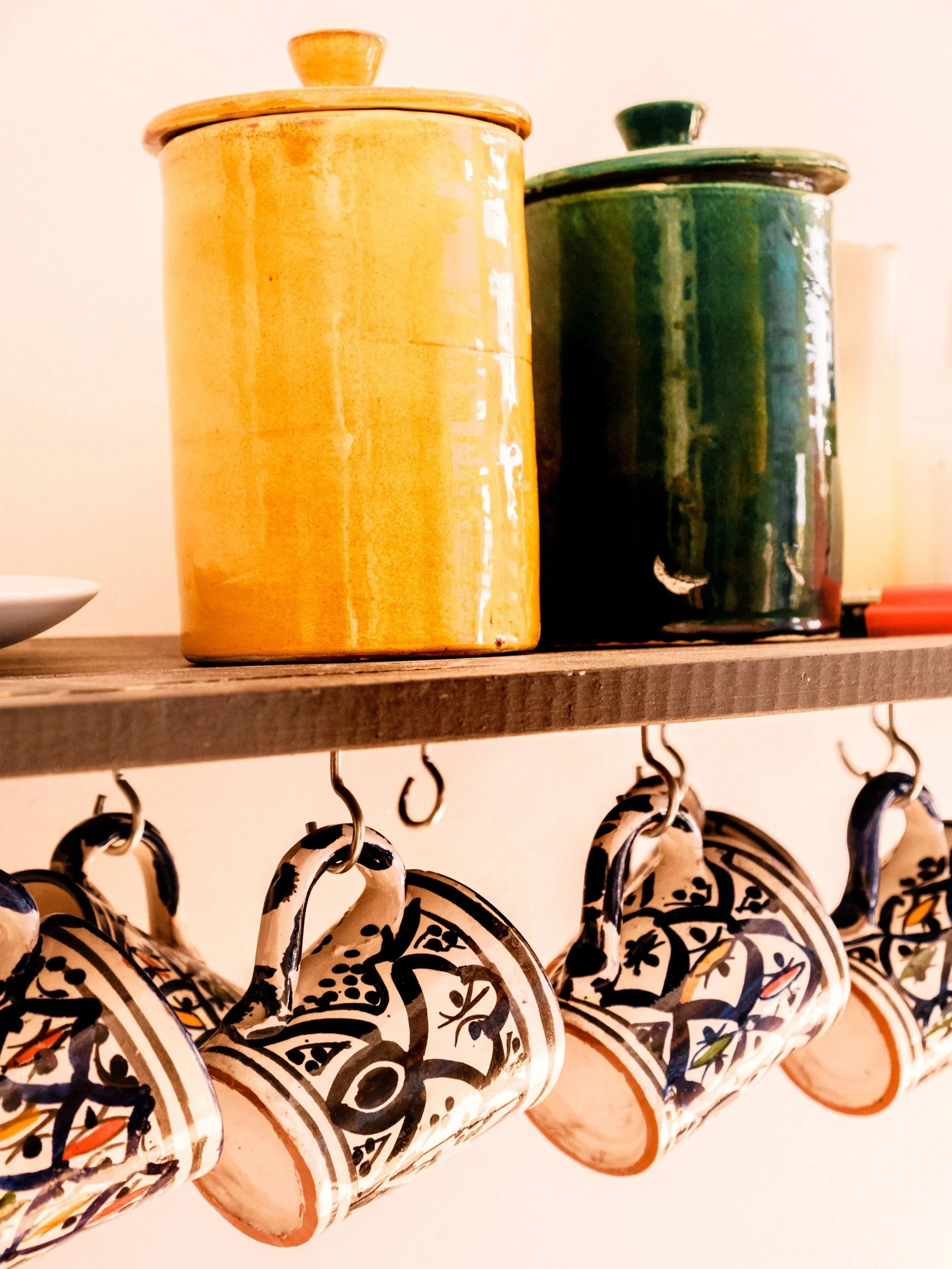 Decorative ceramic mugs hanging from hooks underneath a wooden shelf, with two colorful jars or canisters placed on the shelf.