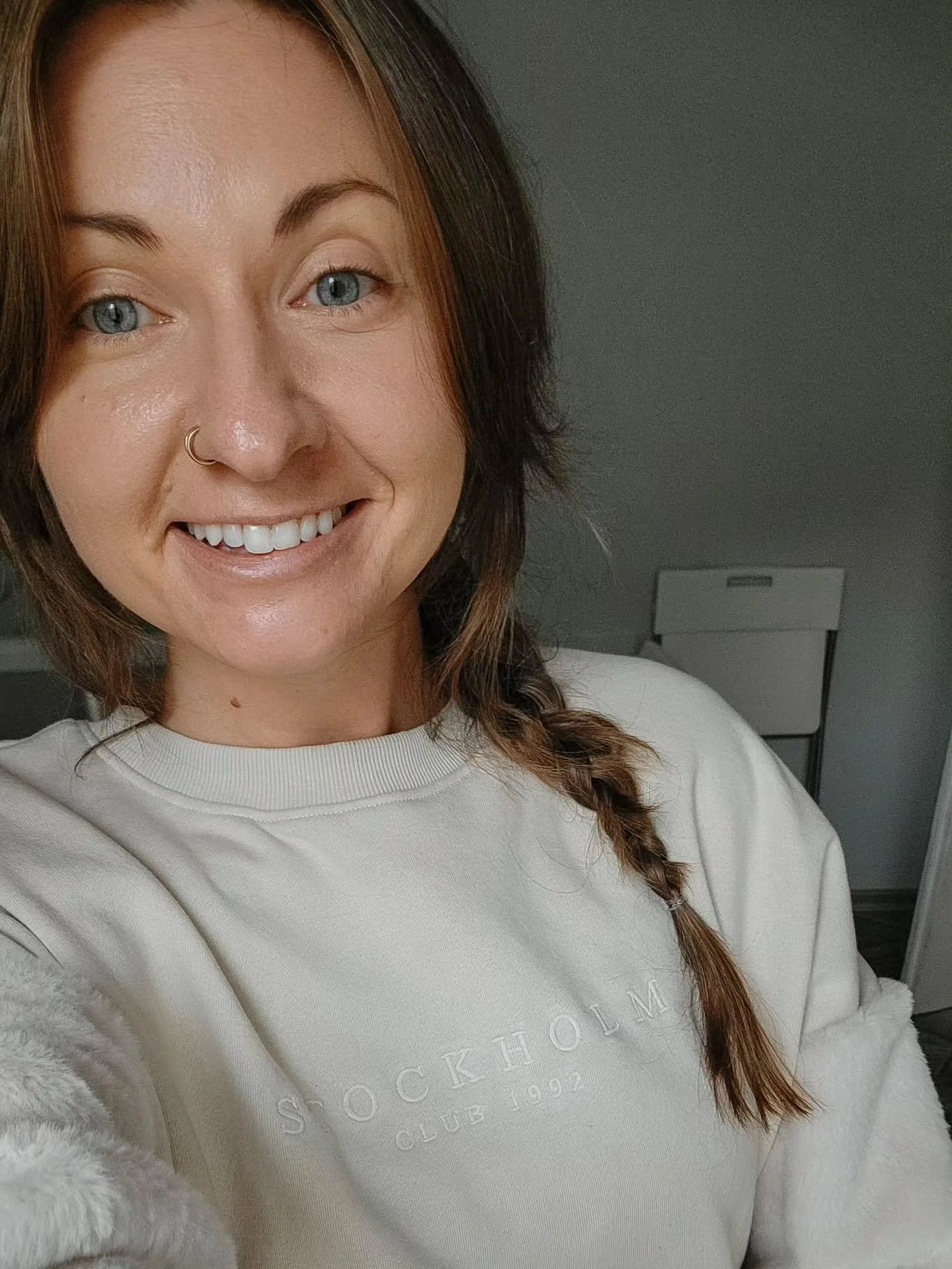 A young woman with light skin, blue eyes, and brown hair with some reddish highlights wearing a cream-colored sweatshirt and a small nose ring, smiling at the camera.