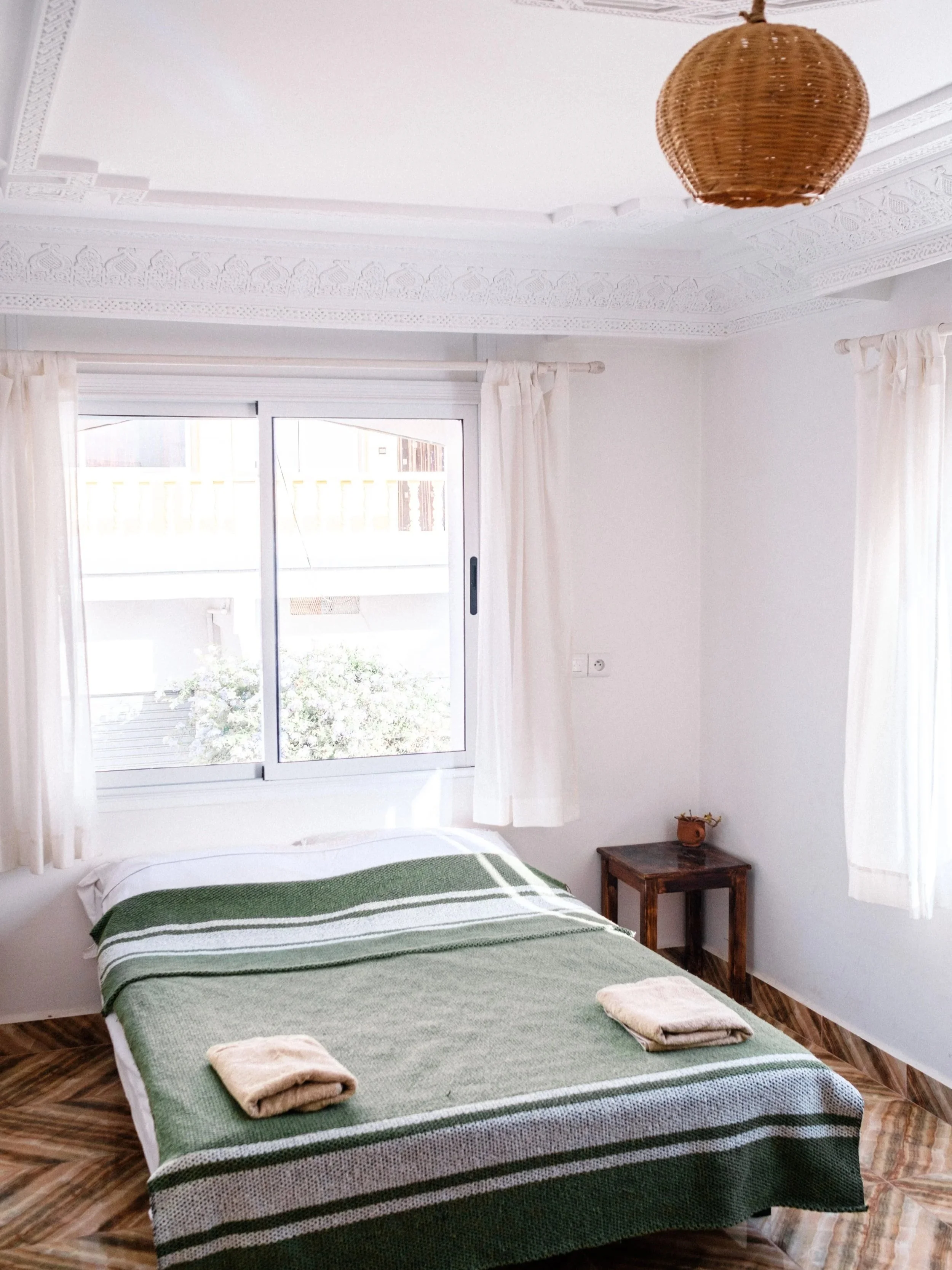 A bedroom with a bed covered in a green and white striped blanket, two folded towels on top, white curtains, a small wooden side table with a plant, a window with a view of outside, and a wicker ceiling light.