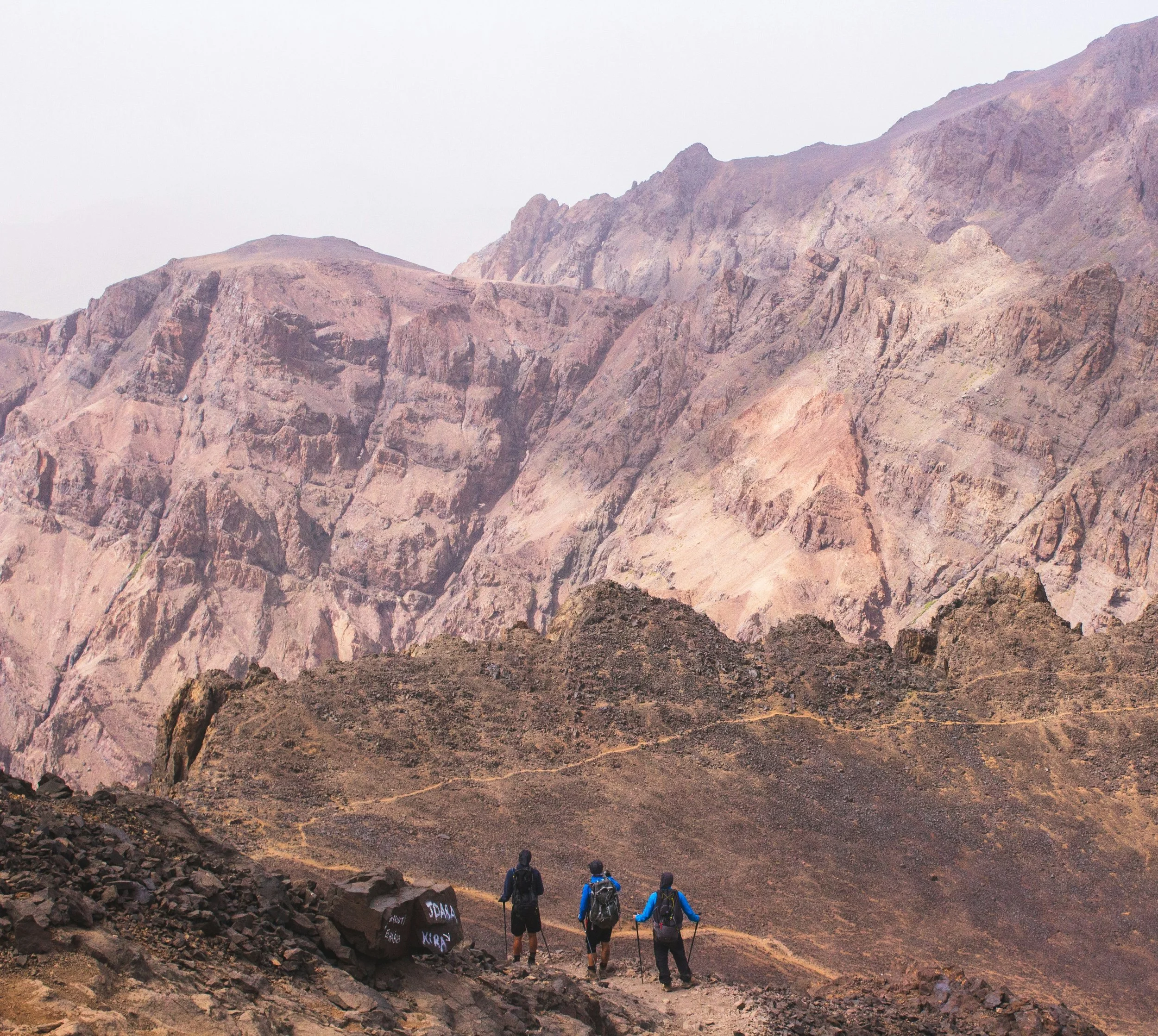 Three hikers walking on a trail in a mountainous landscape with rugged terrain and steep cliffs in the background.
