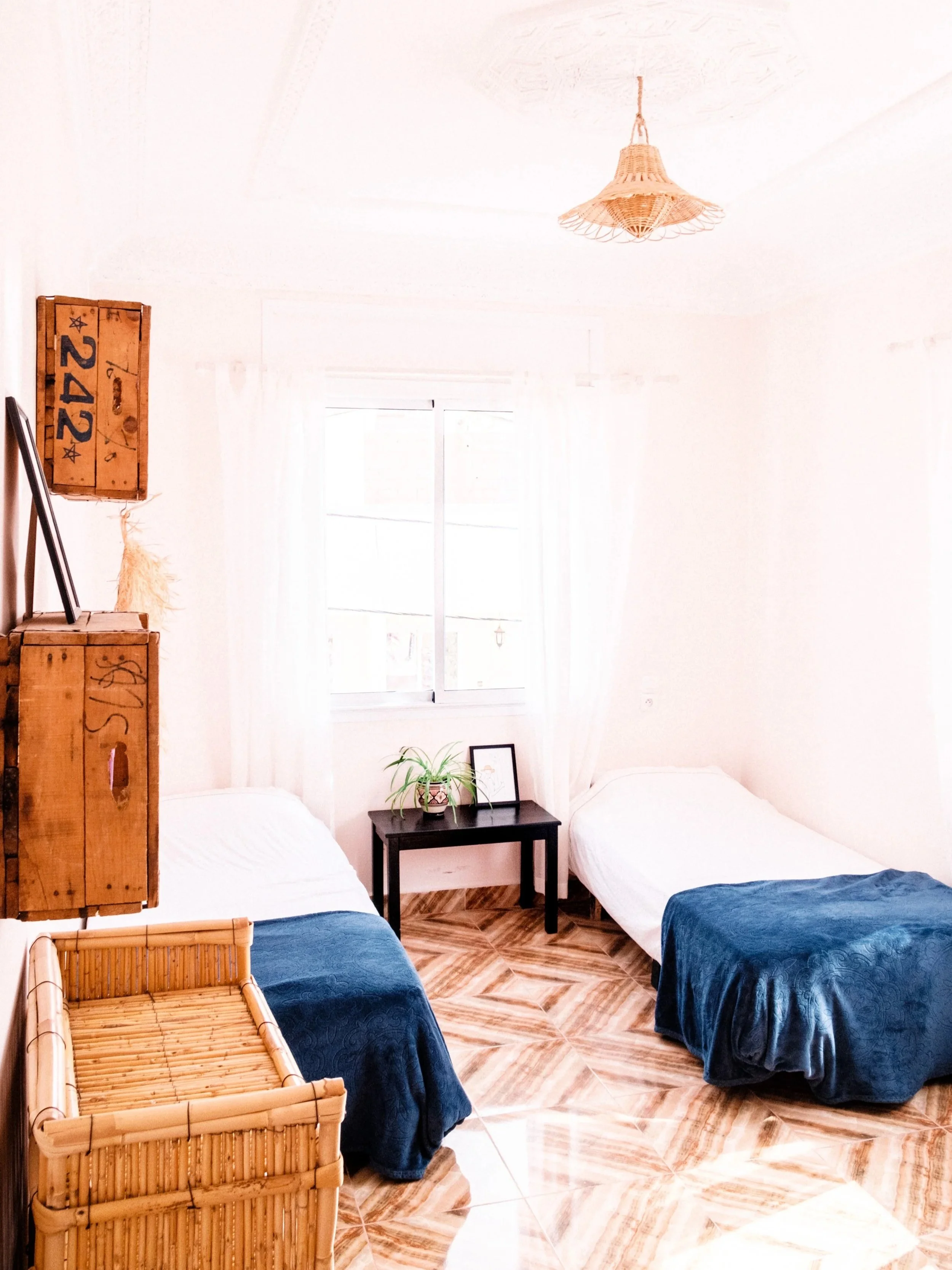 A bright bedroom with two single beds with white sheets and dark blue blankets, a black nightstand with a potted plant and picture frame, a window with sheer curtains, wooden floor tiles, and a wicker light fixture hanging from the ceiling.