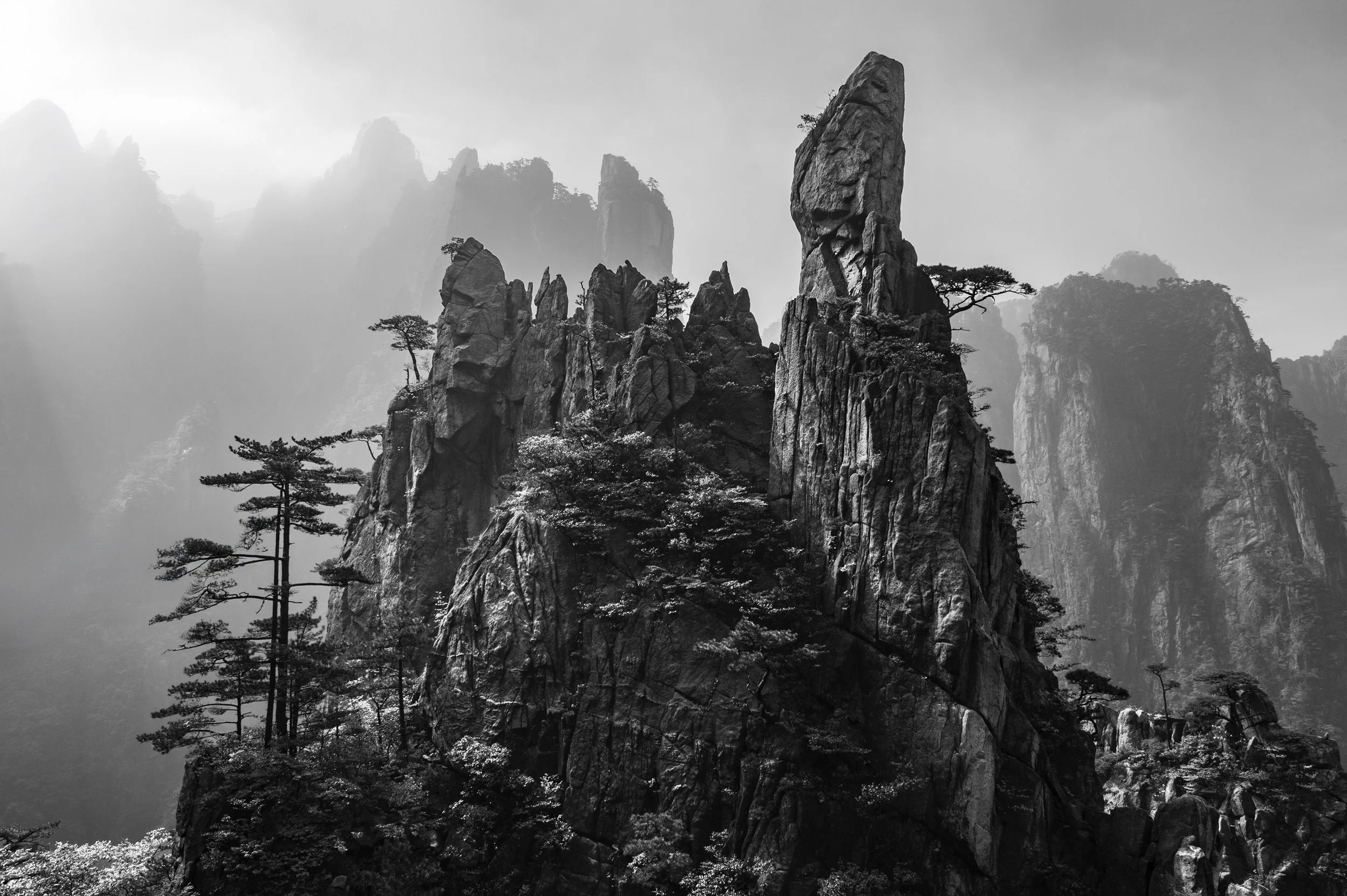 Black and white photo of tall, jagged mountain peaks with sparse trees on the rocky slopes.