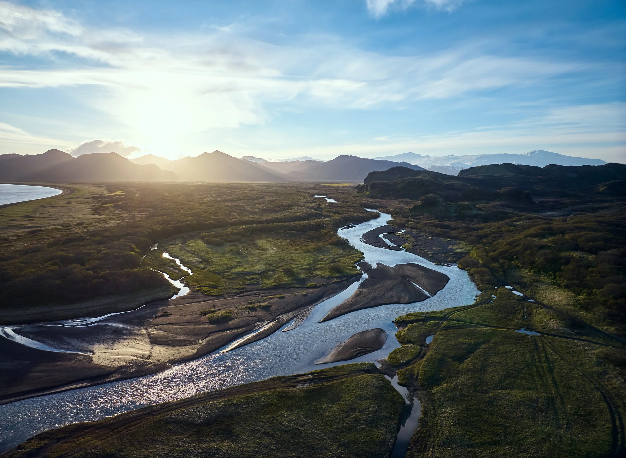 Aerial view of a winding river flowing through green fields with mountains in the background at sunset or sunrise.