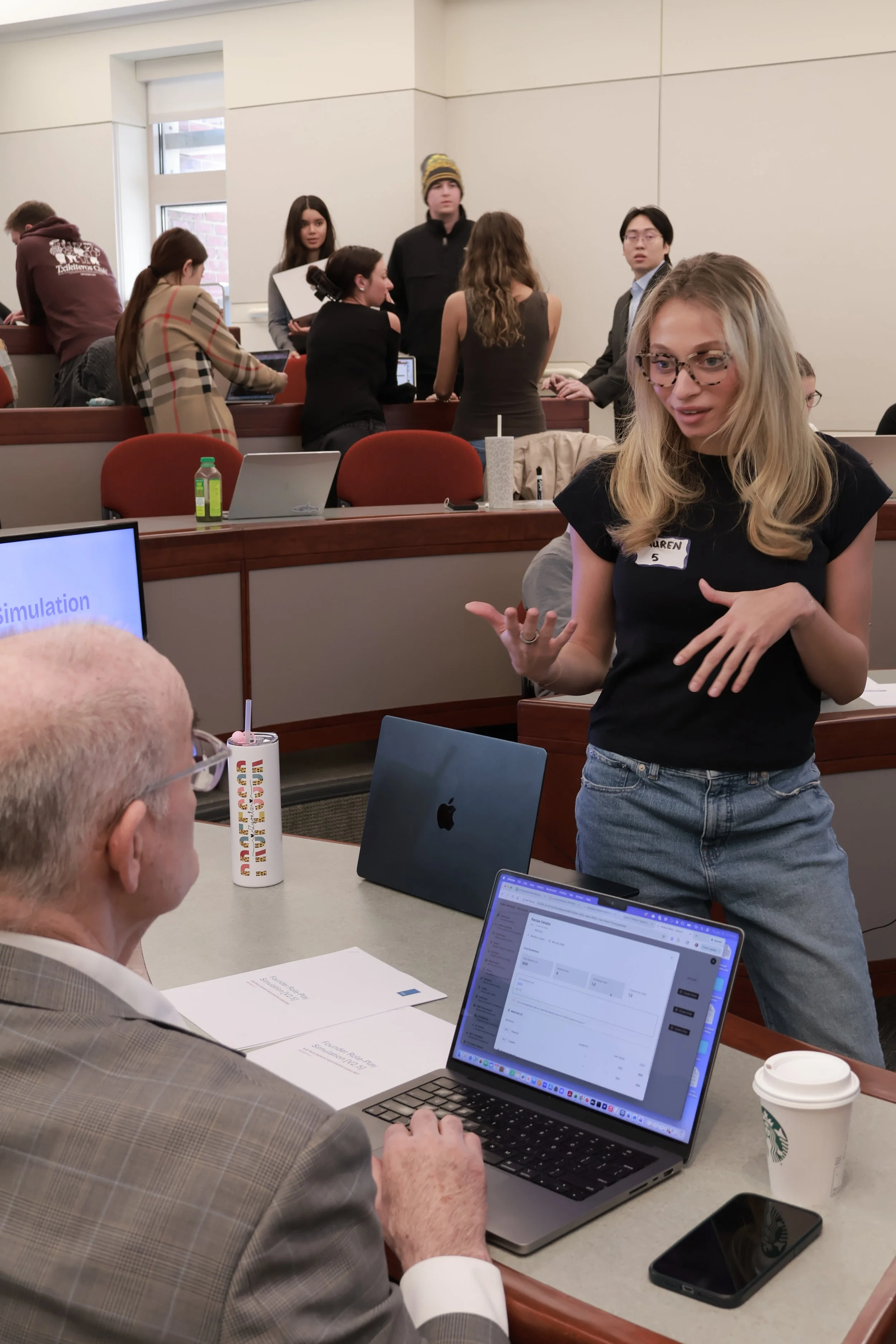 A professor evaluating and scoring a student's live presentation during a SimBus business simulation at Boston University.