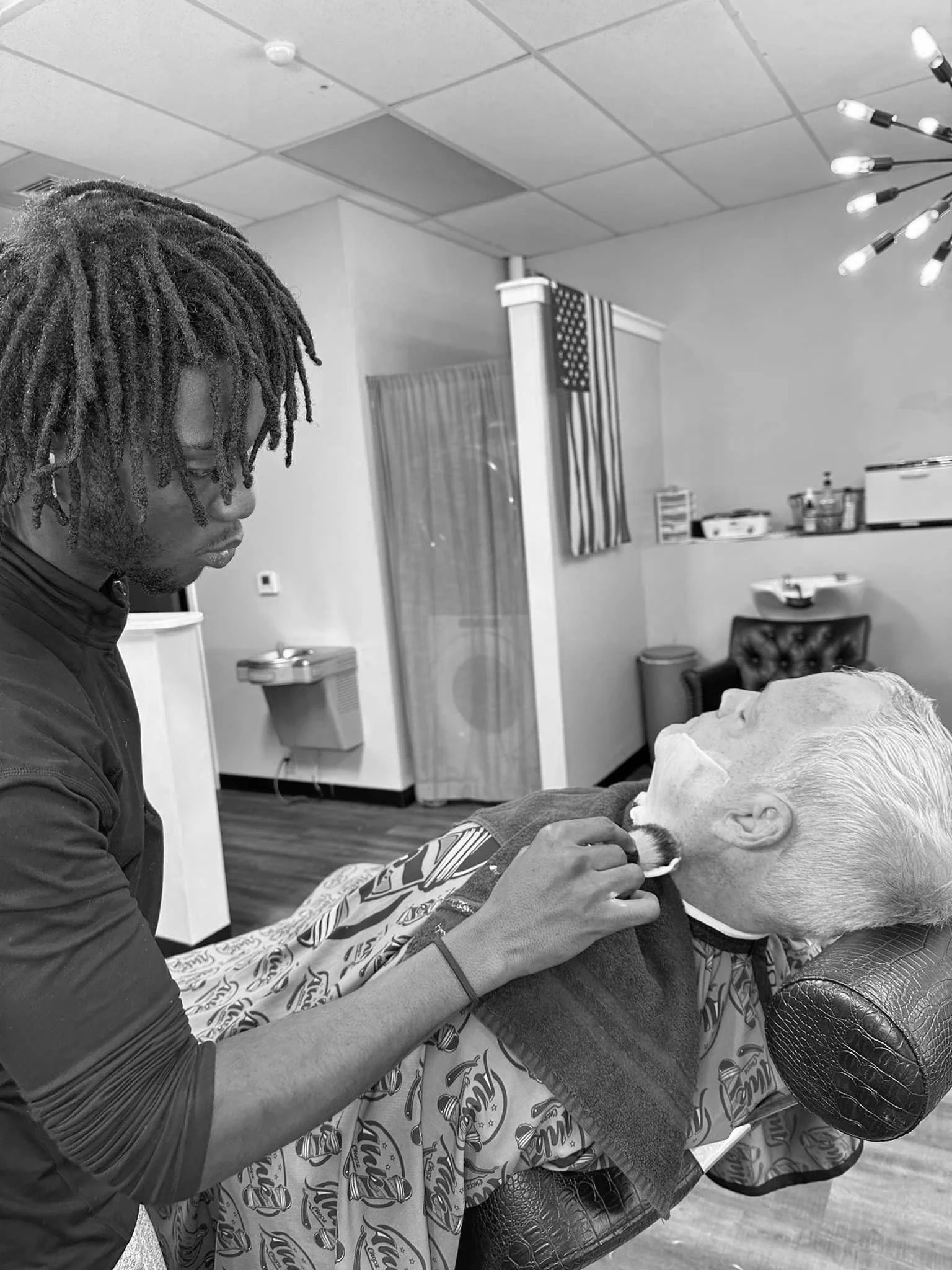 A person getting a beard trim at a barber shop, with the barber using clippers on the client's chin, who is reclined in a barber chair with a patterned cape, in black and white.