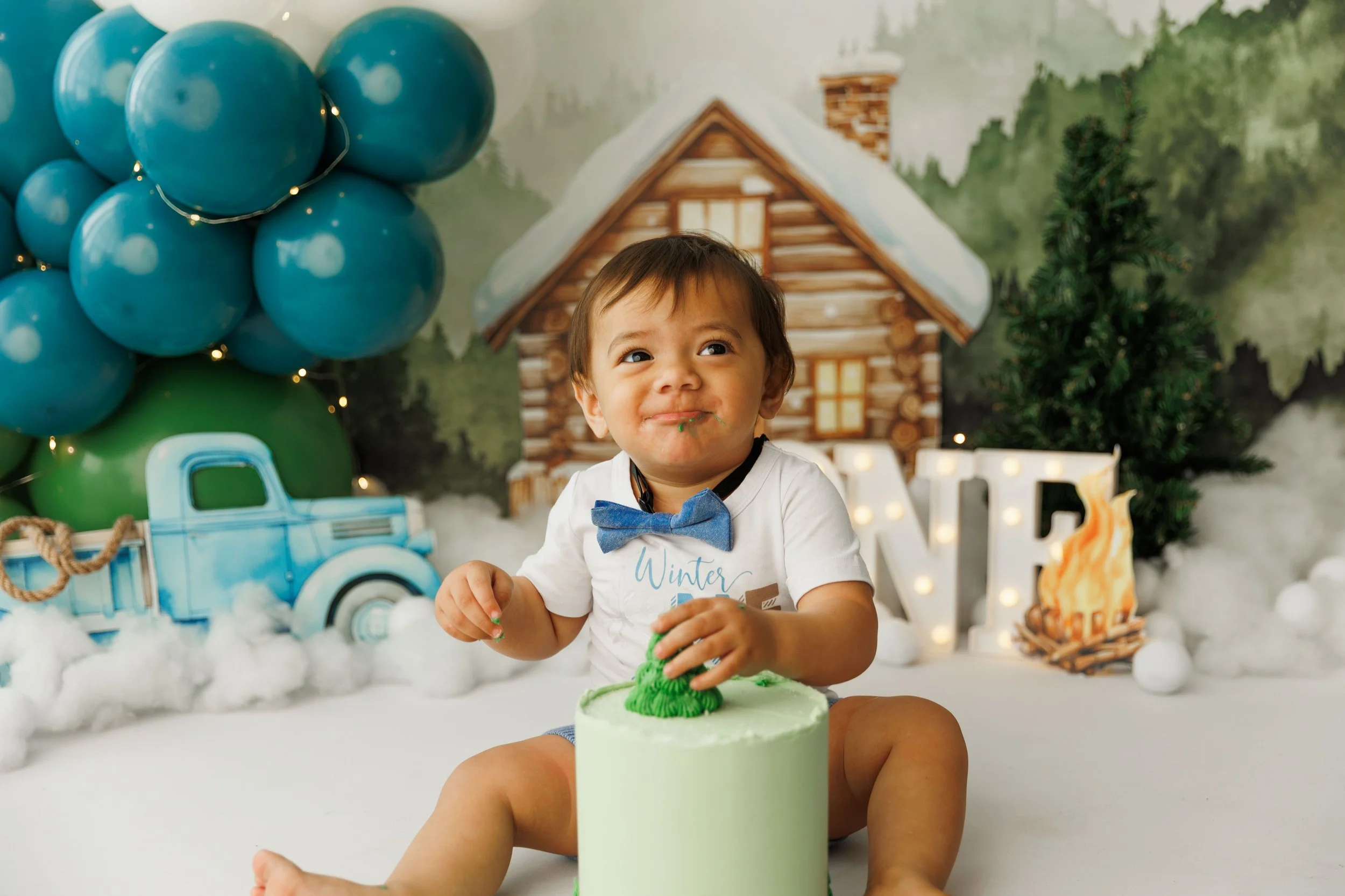 A young child sitting in front of a birthday cake with green frosting, decorated with green and white elements, during a winter-themed celebration. The child is smiling and has some green frosting on their mouth. The background features a painted scene of a log cabin with snow and greenery, along with blue and green balloons, a model truck, and decorative lights.