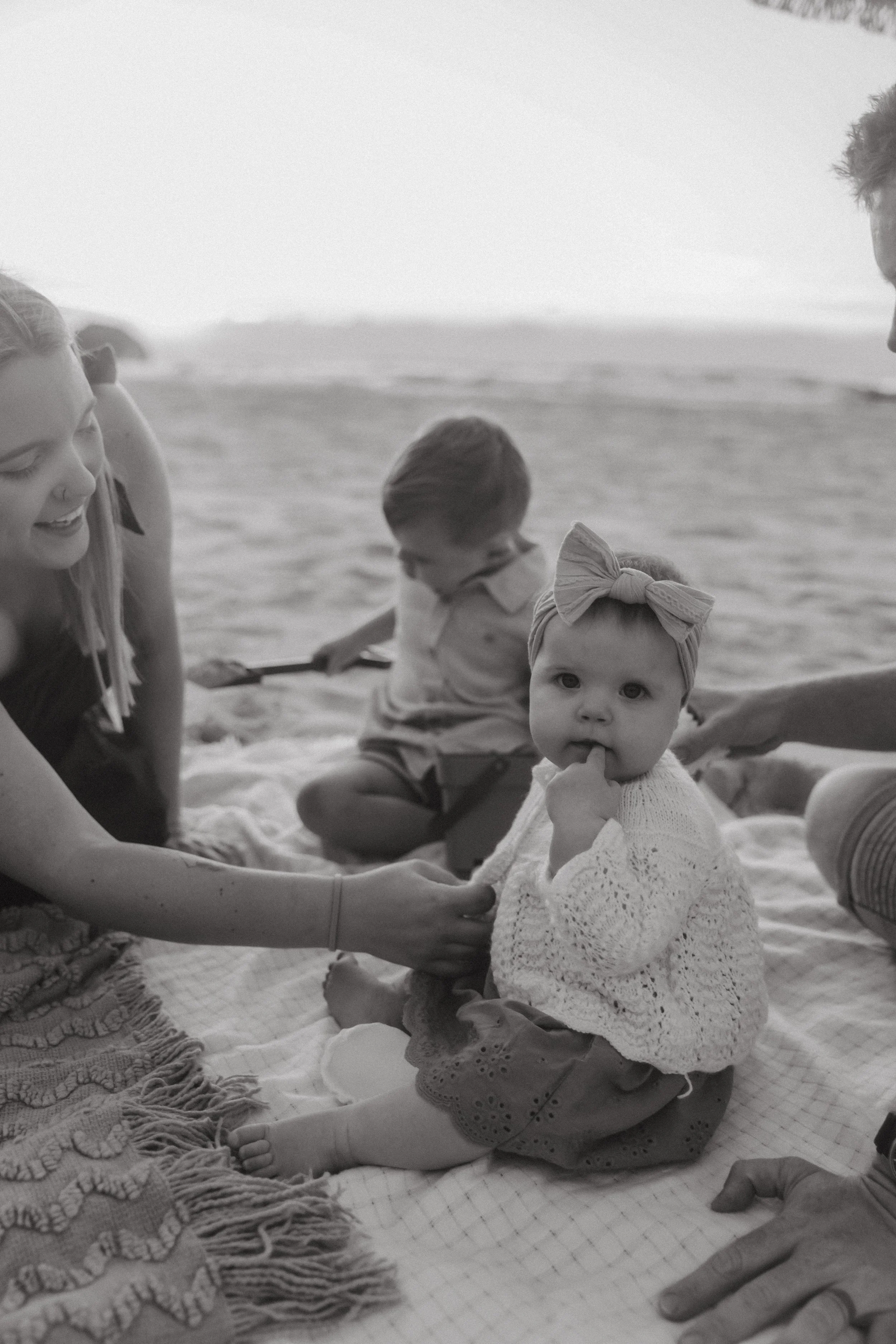 A woman and two children sitting on a blanket at the beach, with ocean waves in the background. The girl has a headband and is touching her mouth, while a boy is looking down and a baby girl is eating or sucking on her hand.