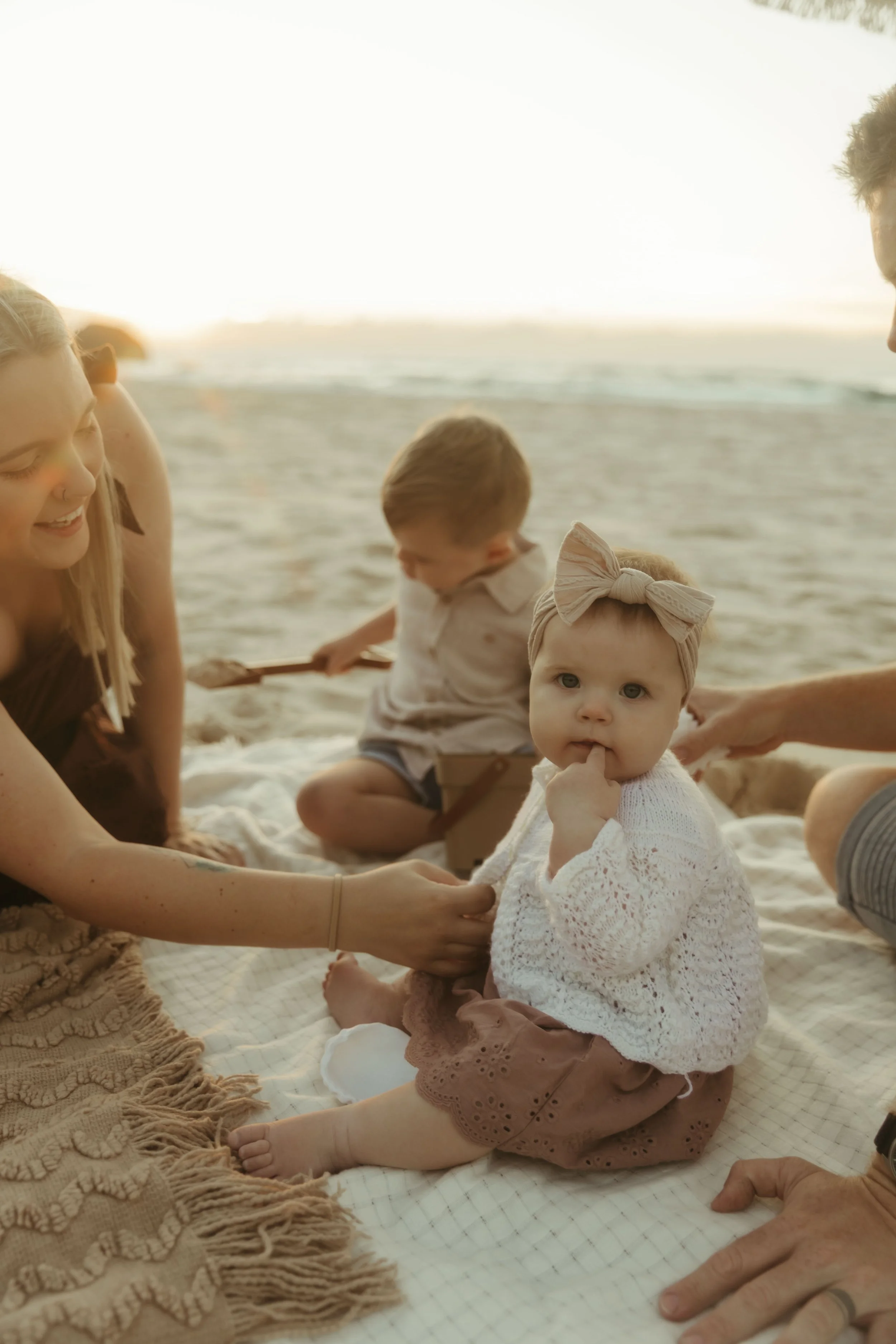 A group of people, including children and adults, sitting on a blanket at the beach during sunset, enjoying and playing with each other.
