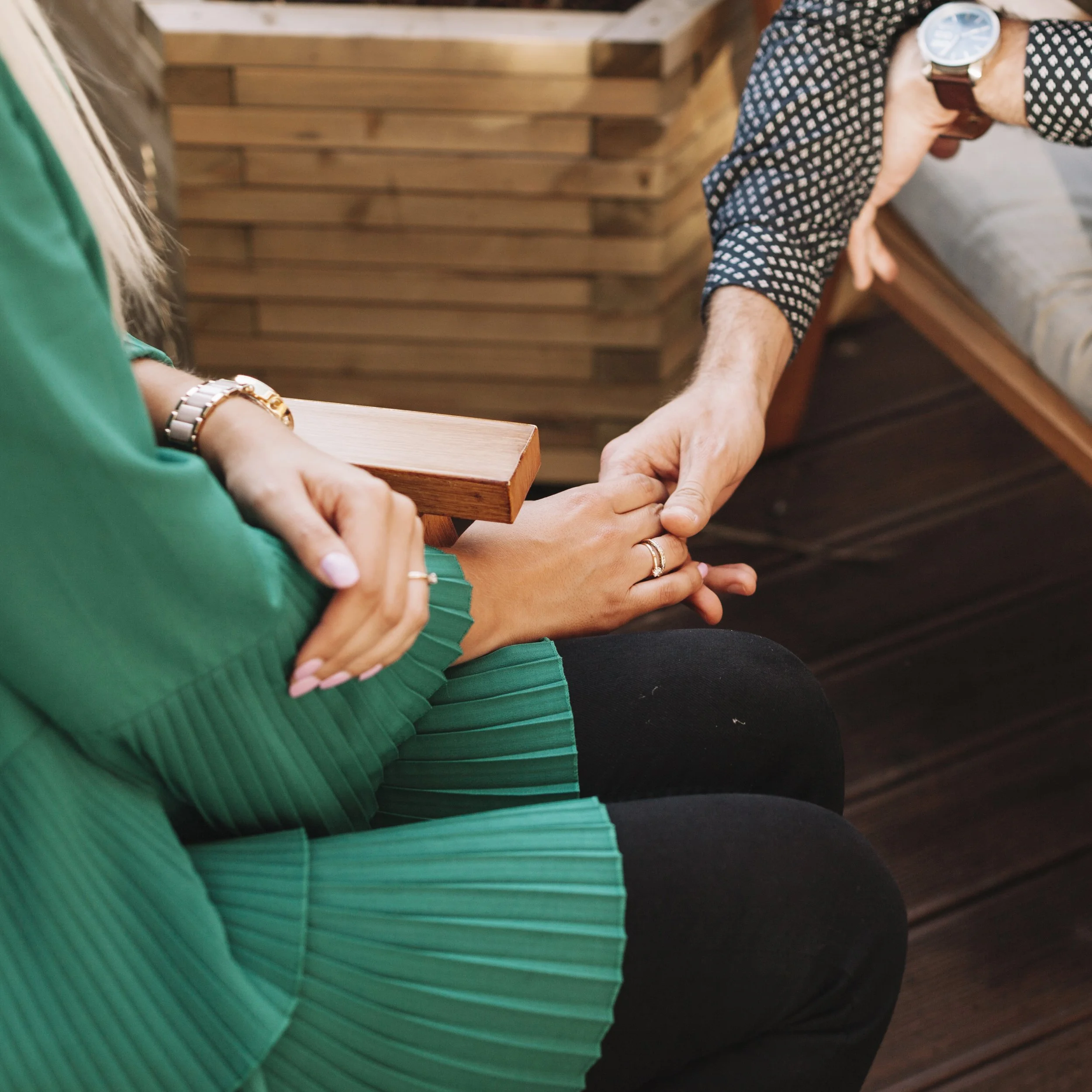 A person in a green pleated skirt and matching top holding hands with another person wearing a checkered shirt, sitting on wooden benches, with their hands clasped and fingers intertwined, in a cozy indoor setting.