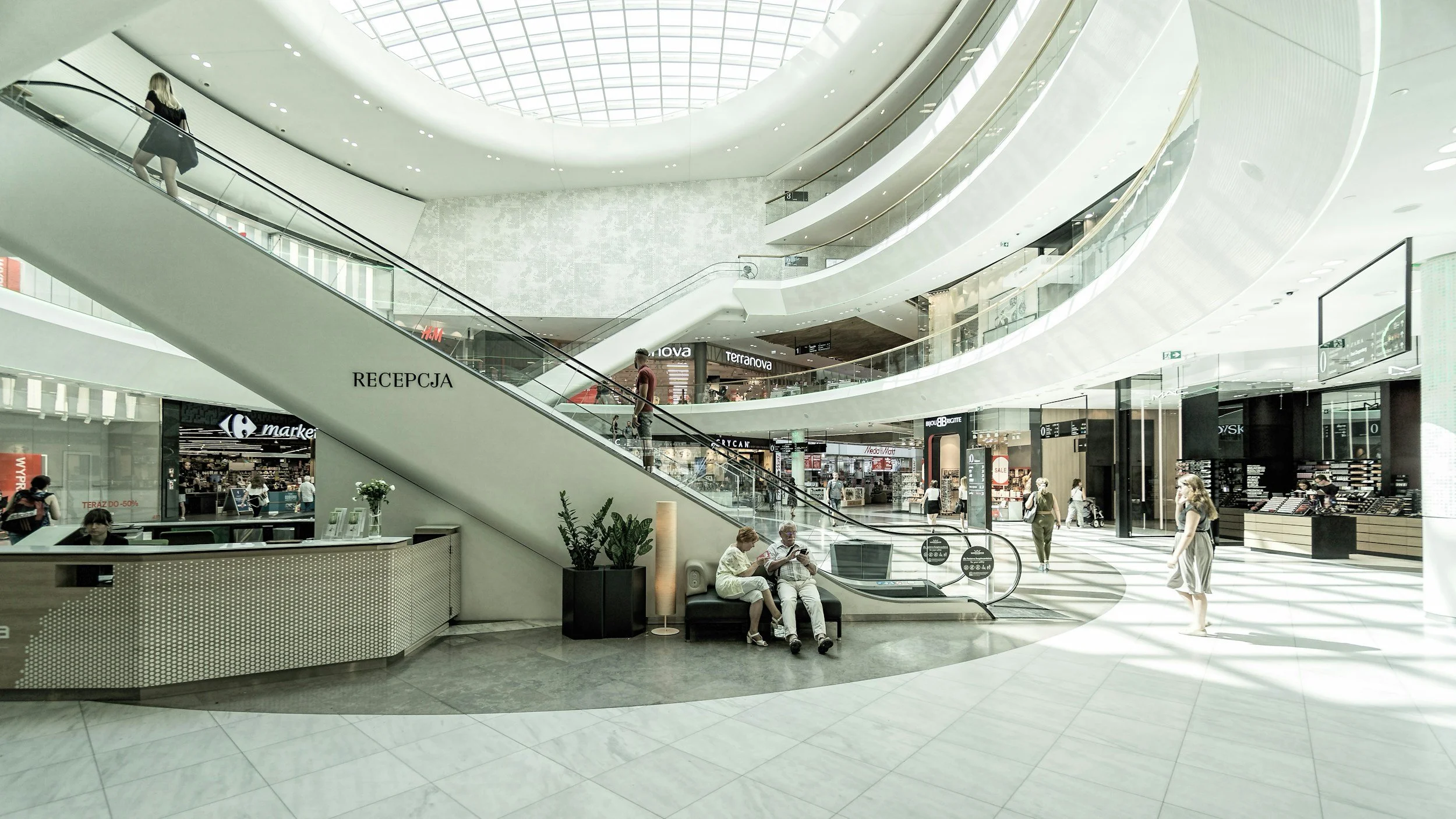 Innenansicht eines modernen Einkaufszentrums mit mehreren Geschossen, Rolltreppen, Passanten und Geschäften.