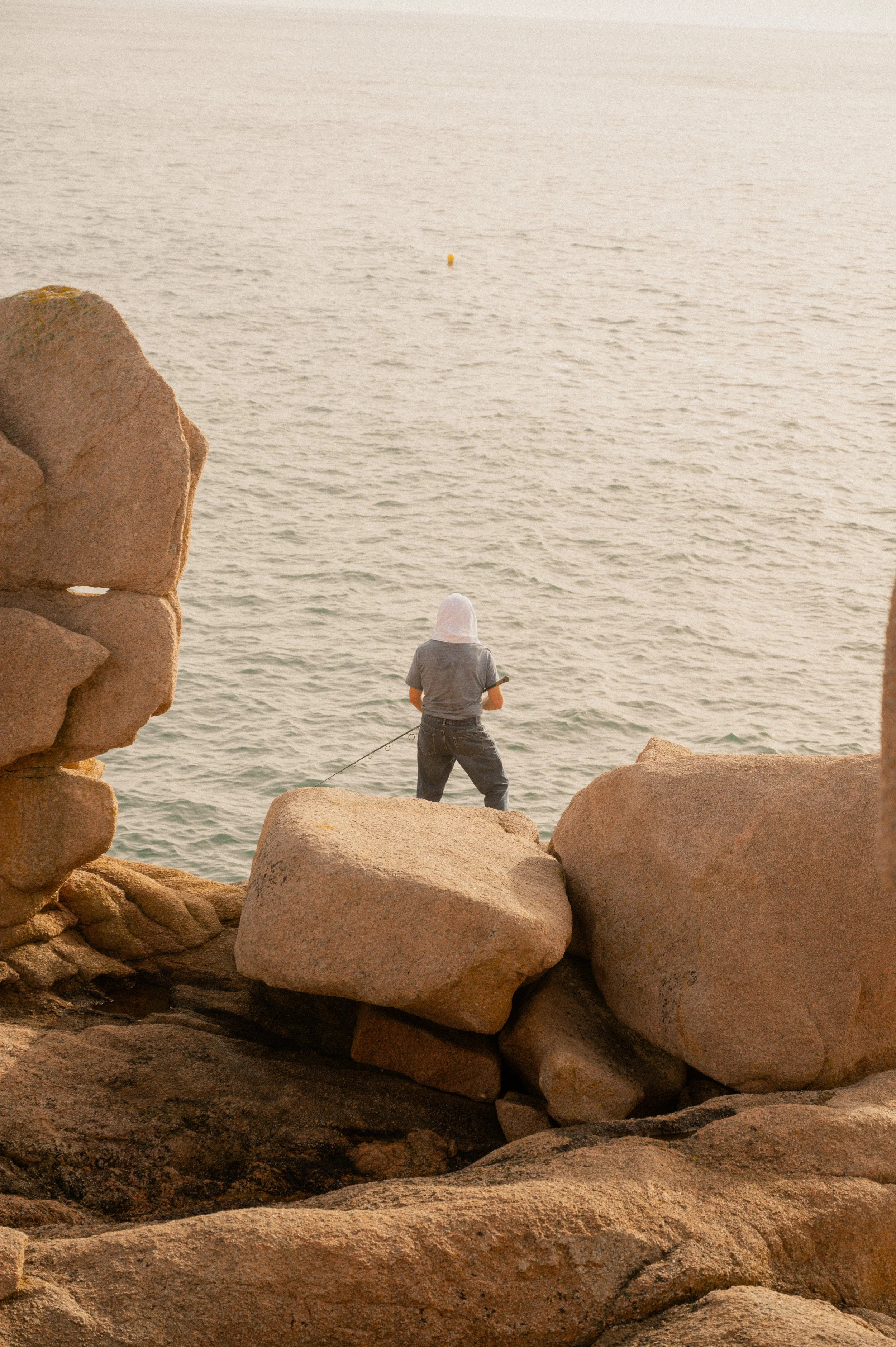 Personne debout sur des rochers au bord de la mer, regardant au loin, en train de pêcher, avec une écharpe blanche couvrant la tête.