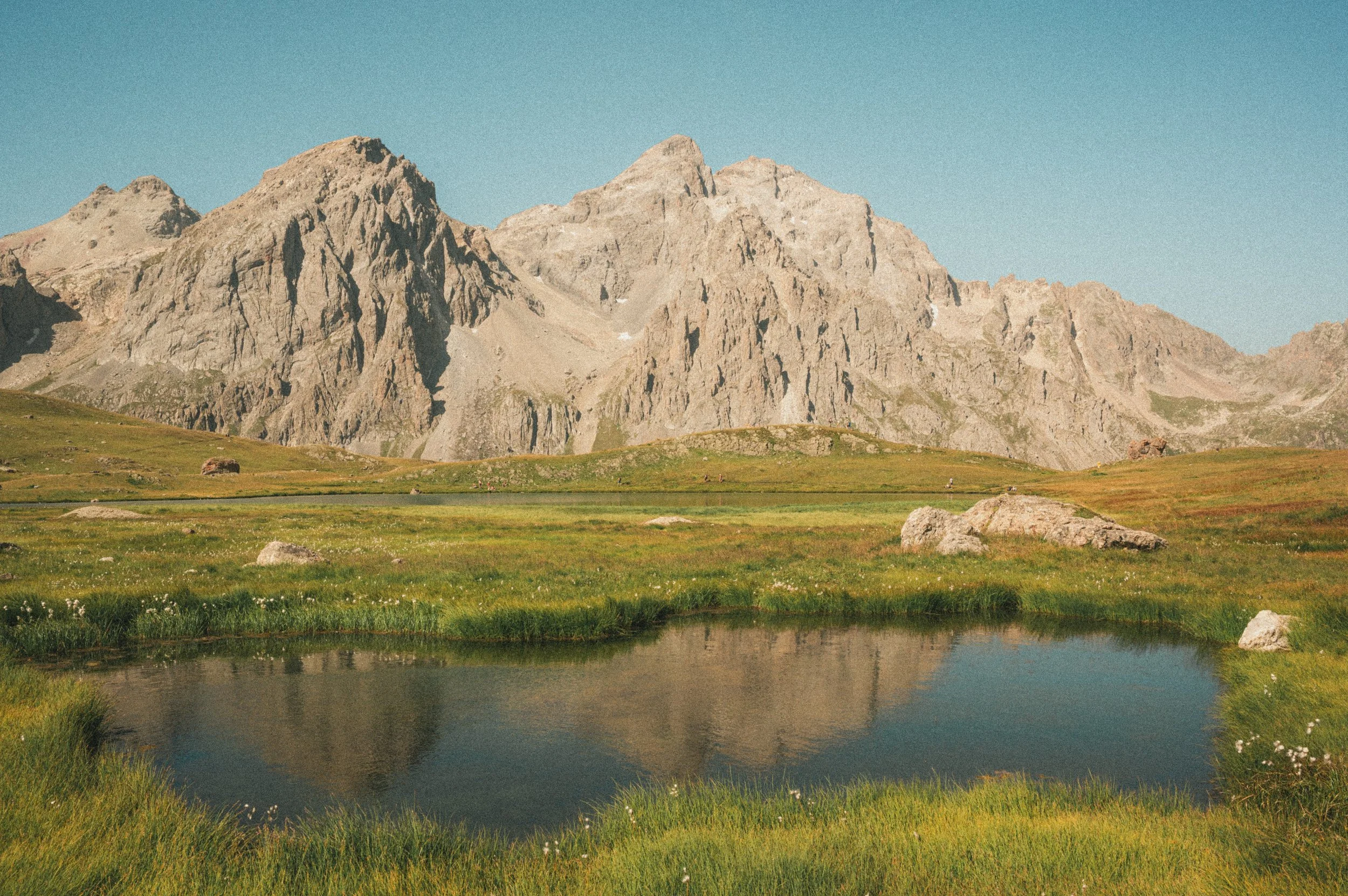 Paysage de montagnes rocheuses avec un lac au premier plan, herbes vertes et un ciel bleu clair.
