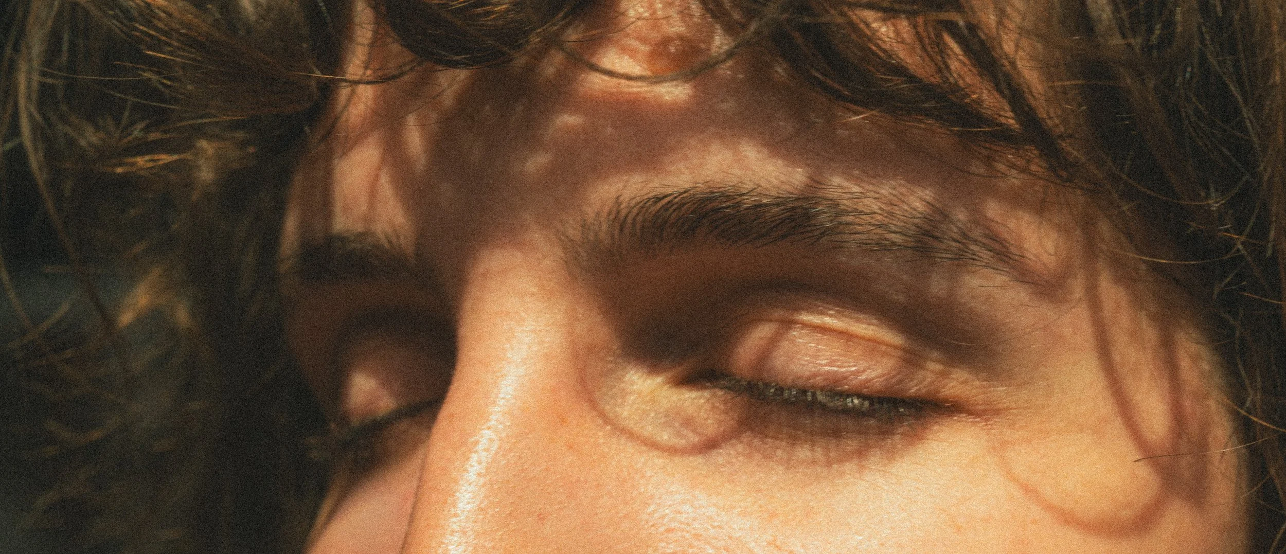 Zoom sur un visage avec les yeux fermés, boucles de cheveux brunes, peau claire, détails des cils et des sourcils.