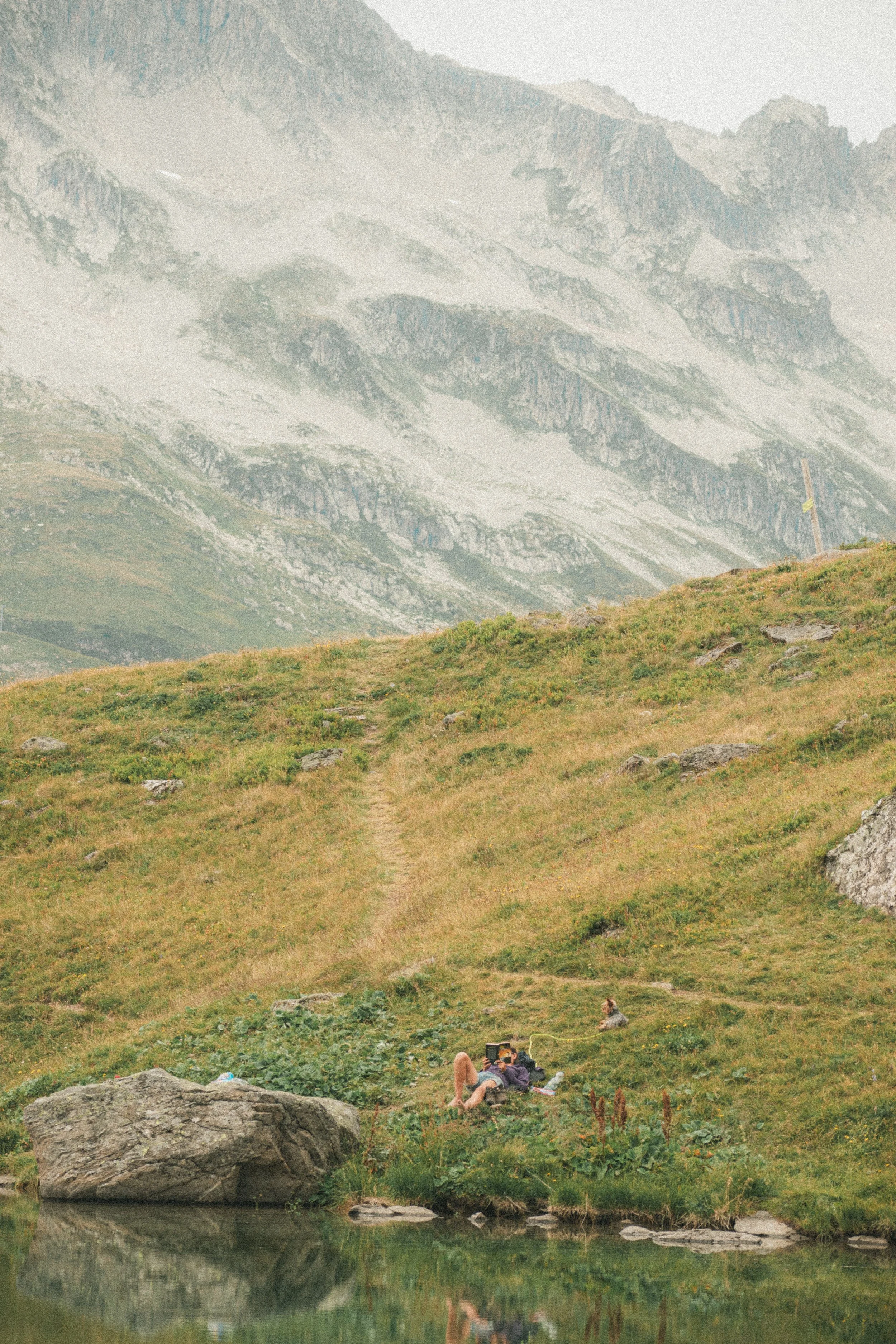 Une personne allongée sur l'herbe au bord d'un étang, prenant une photo avec un téléphone, entourée de rochers et de végétation montagnarde, avec des montagnes enneigées en arrière-plan.