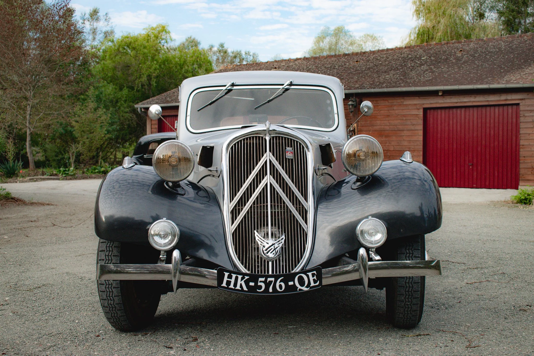 Voiture de rallye rouge équipée de plusieurs phares avant, stationnée sur une surface gravel, avec un bâtiment blanc en arrière-plan.
