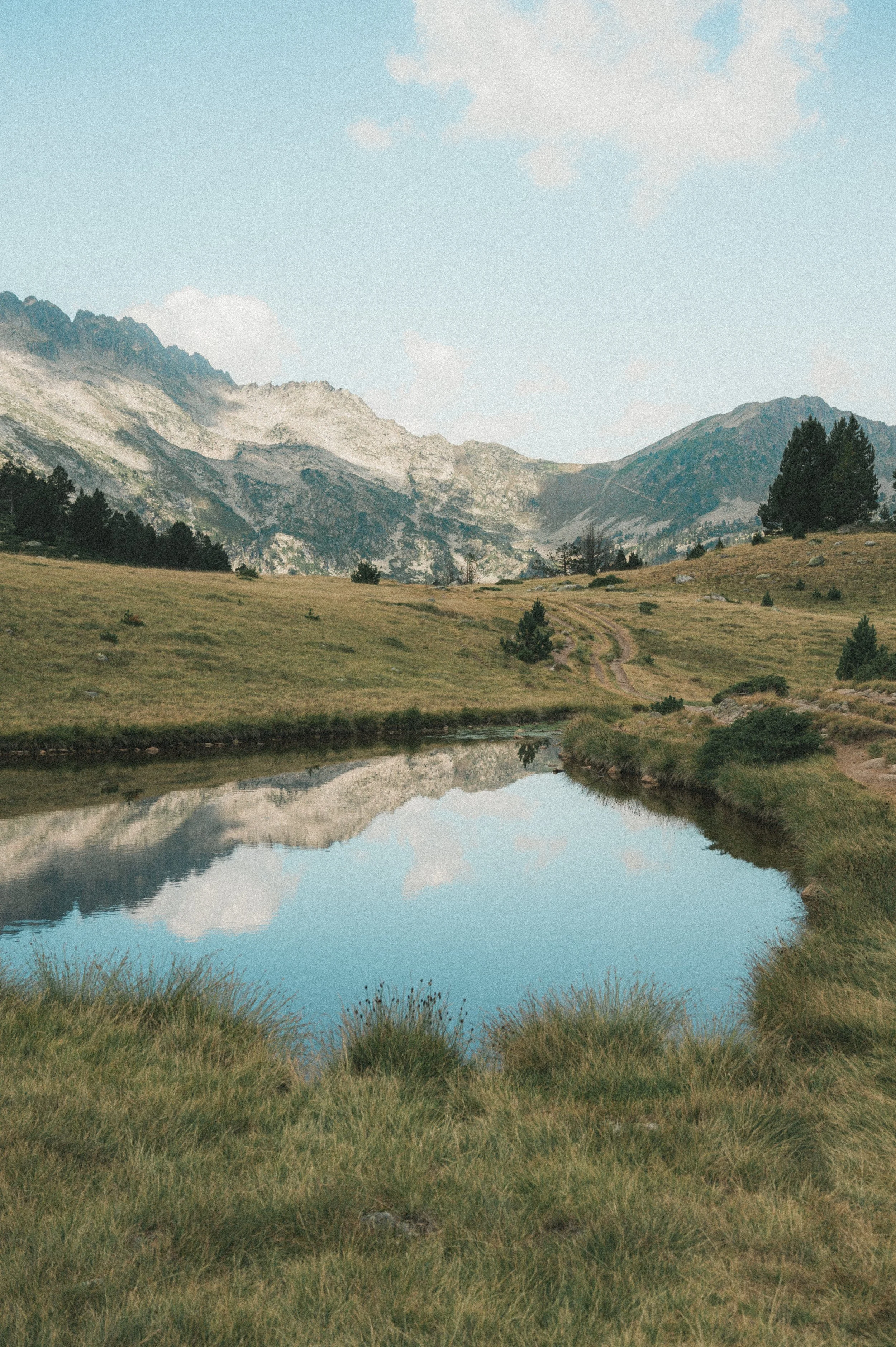 Un paysage naturel avec un lac calme reflétant le ciel bleu, entouré de collines verdoyantes et de montagnes en arrière-plan.