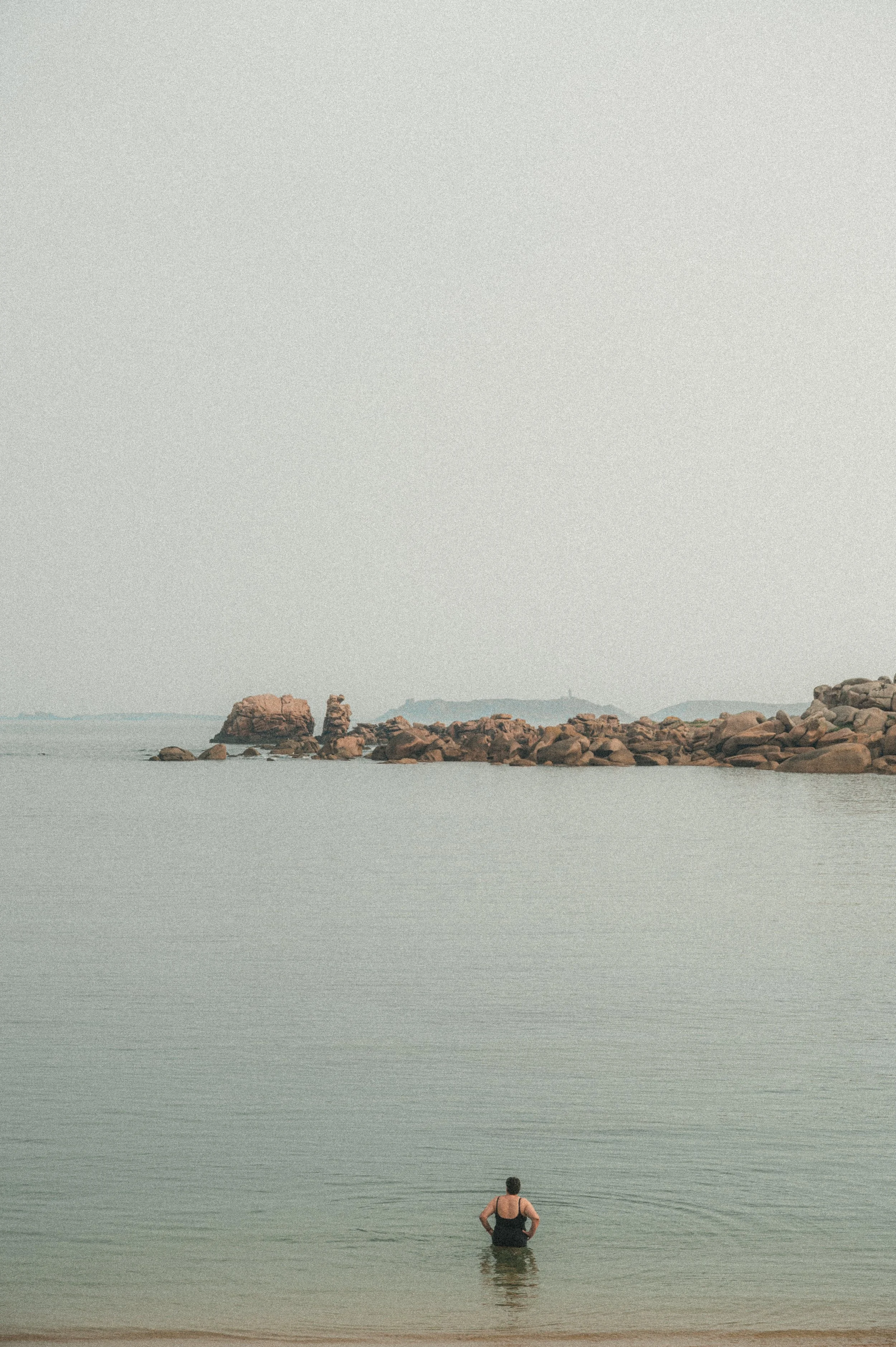 Une personne se tient dans l'eau au bord de la mer, regardant vers des rochers au large sous un ciel clair.
