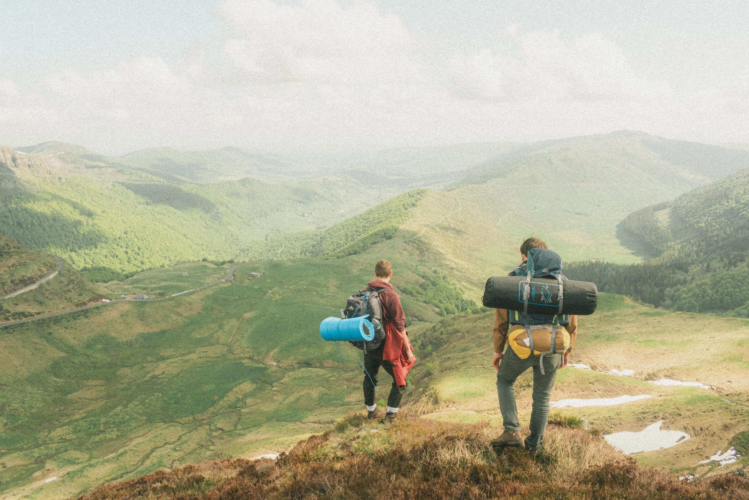 Deux randonneurs avec de grands sacs à dos parcourent une vallée verdoyante avec des collines et des montagnes en arrière-plan.